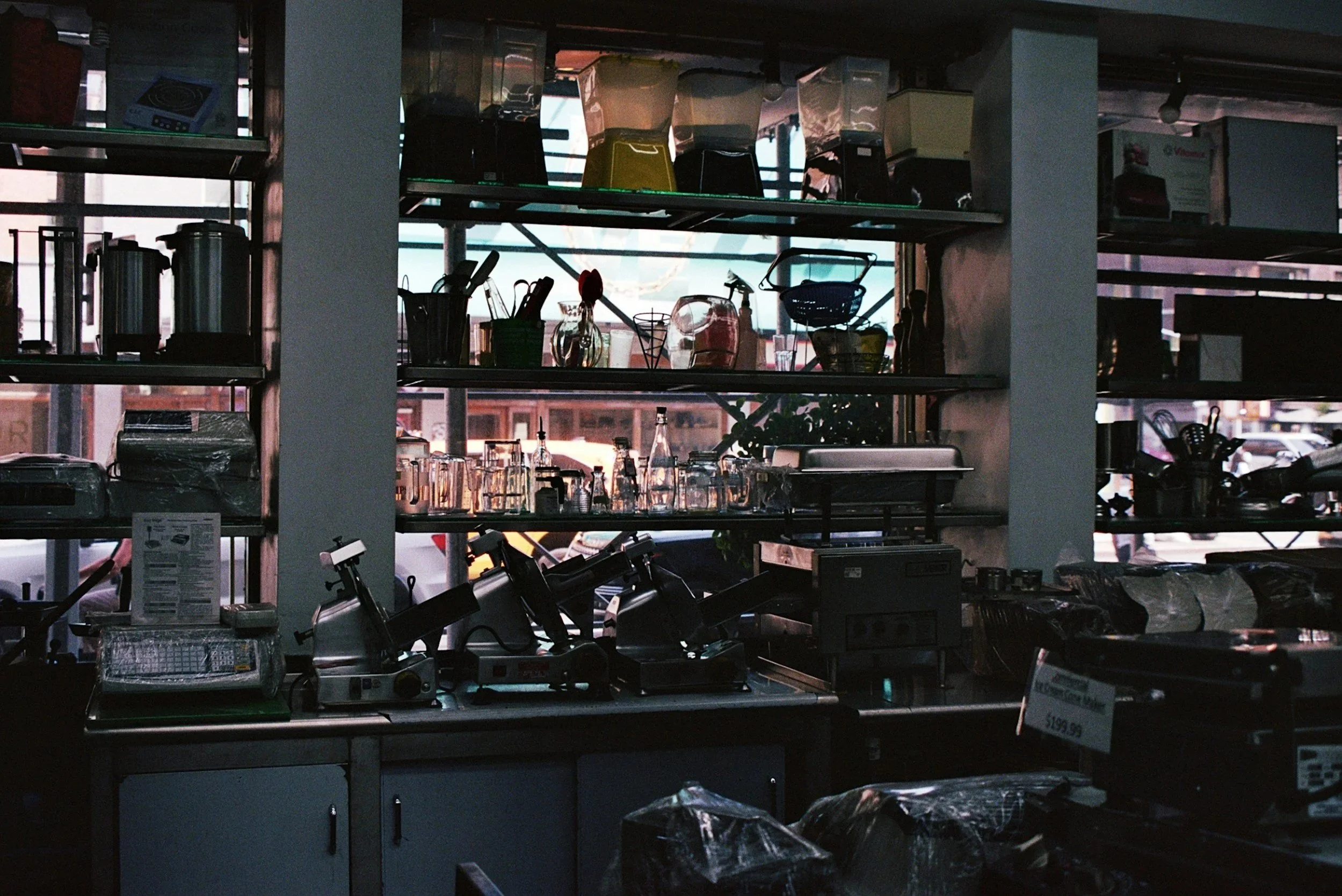 Shelves in a store with kitchen utensils and appliances silhouetted against a window, with some sunlight and outdoor buildings visible in the background.