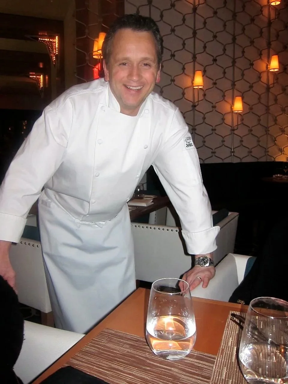 A smiling male chef in a white chef coat leaning over a table in a restaurant, with a glass of water in front of him and warm lighting in the background.