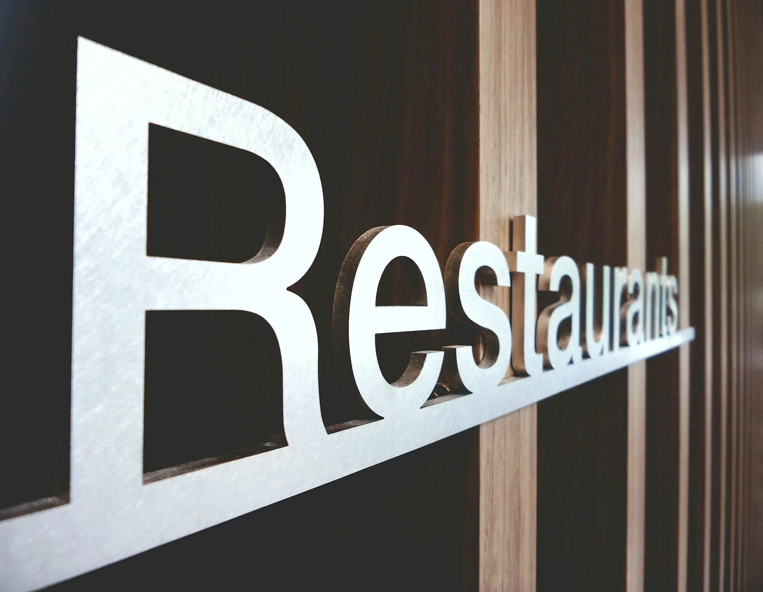 Close-up of three-dimensional white letters on a black background, spelling out part of a word, with a wooden wall in the background.