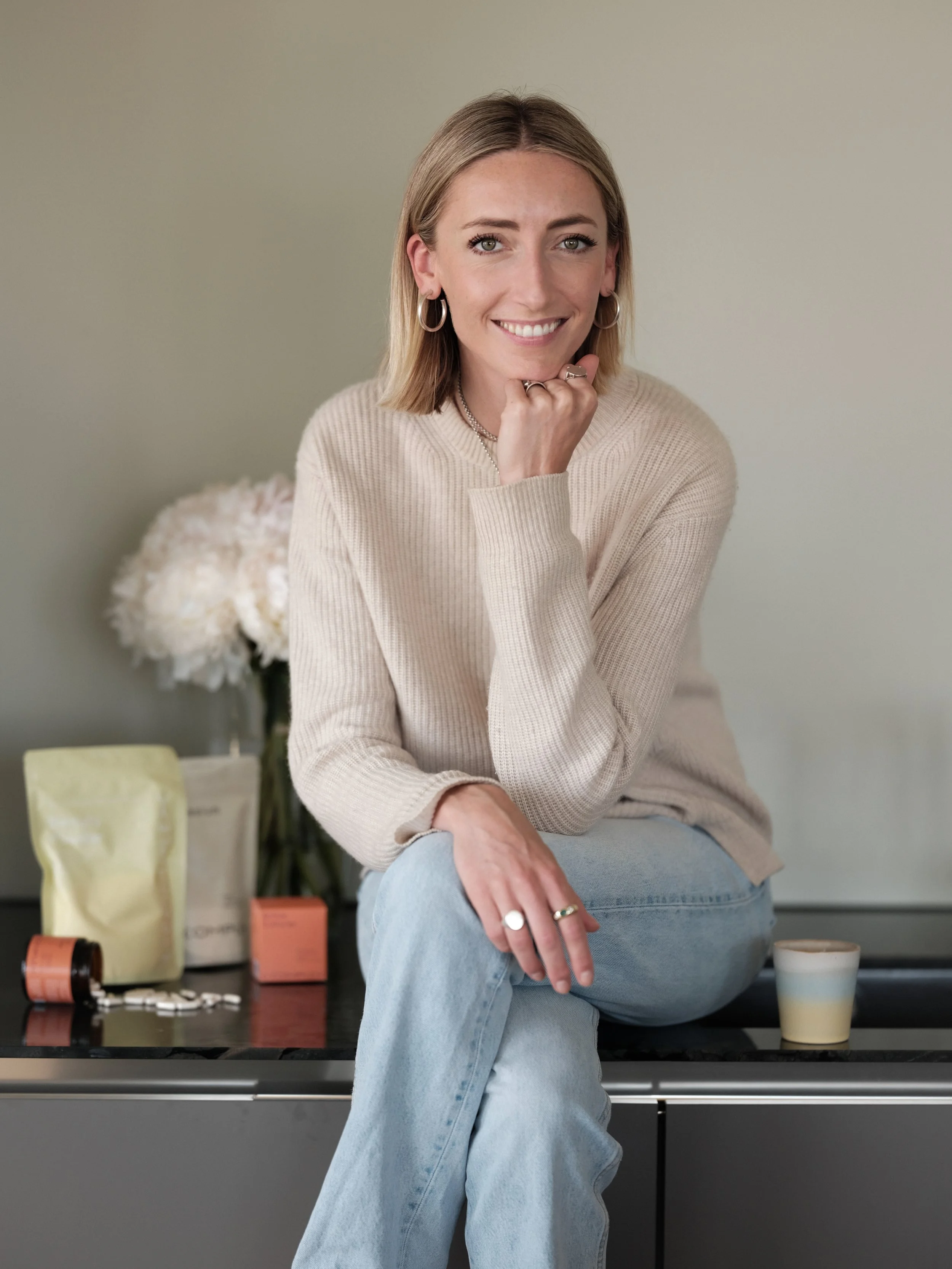 A woman with shoulder-length blonde hair, wearing hoop earrings, a beige sweater, and light blue jeans, sitting on a black countertop with her hand resting on her chin and smiling at the camera. Behind her, there are various bottles and containers, a vase with white flowers, and a yellow bag with a white object inside.