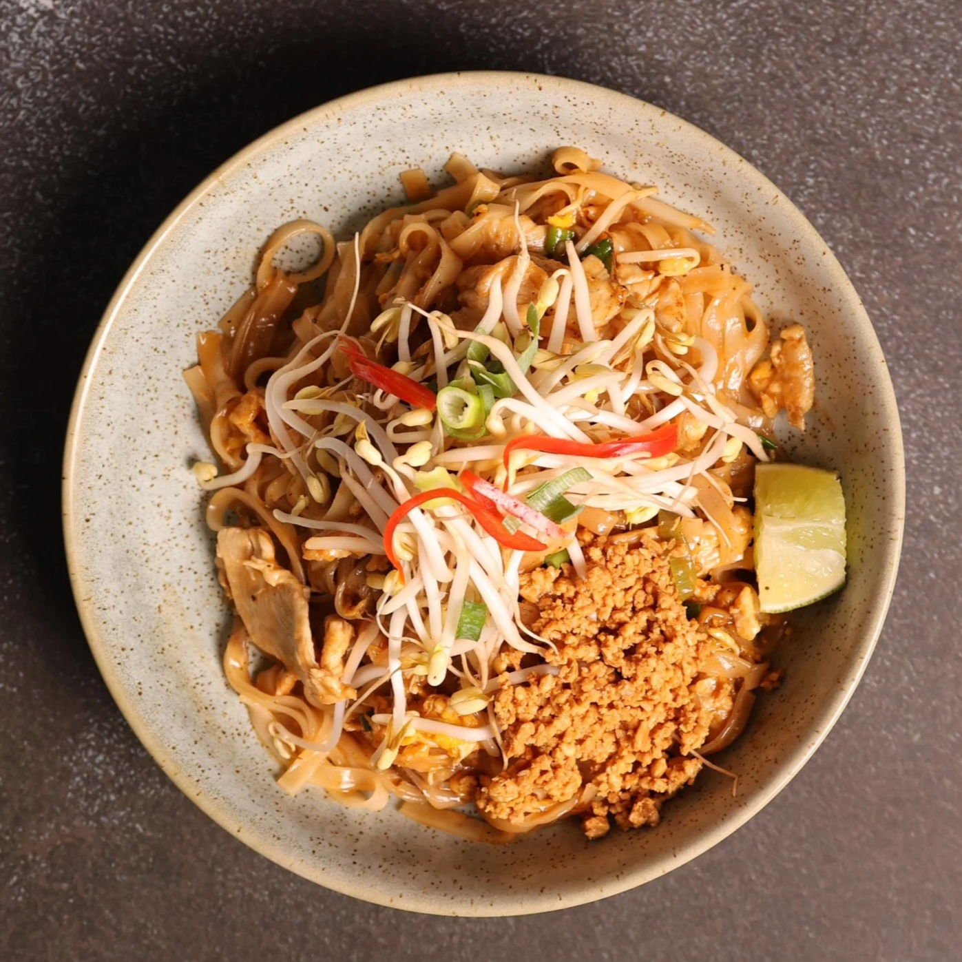 A bowl of Asian stir-fried noodles topped with bean sprouts, chopped green onions, sliced red chili, ground peanuts, and a lime wedge on the side.