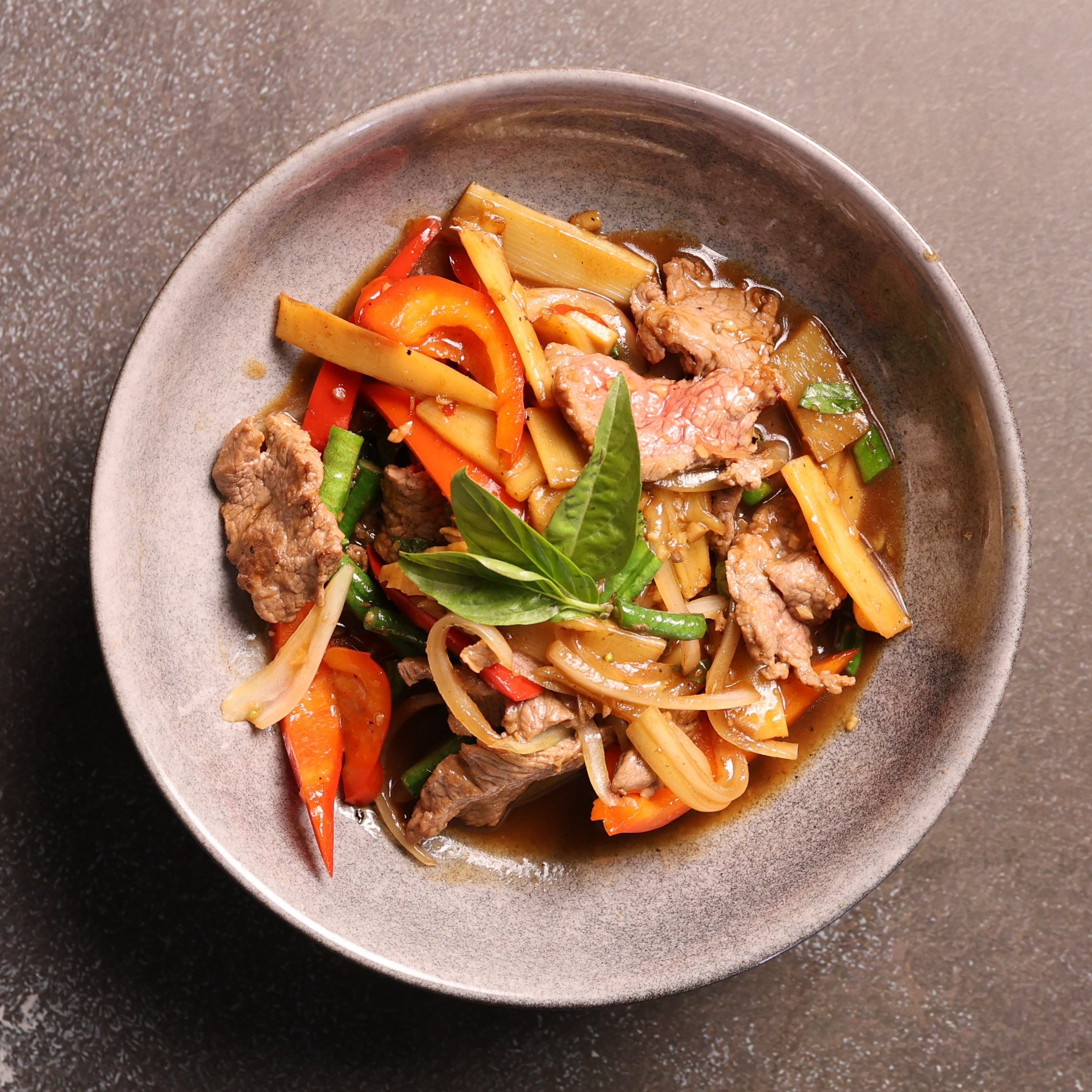 A bowl of Asian-style noodle soup with slices of beef, red bell peppers, green onions, bamboo shoots, and basil leaves in a savory broth.
