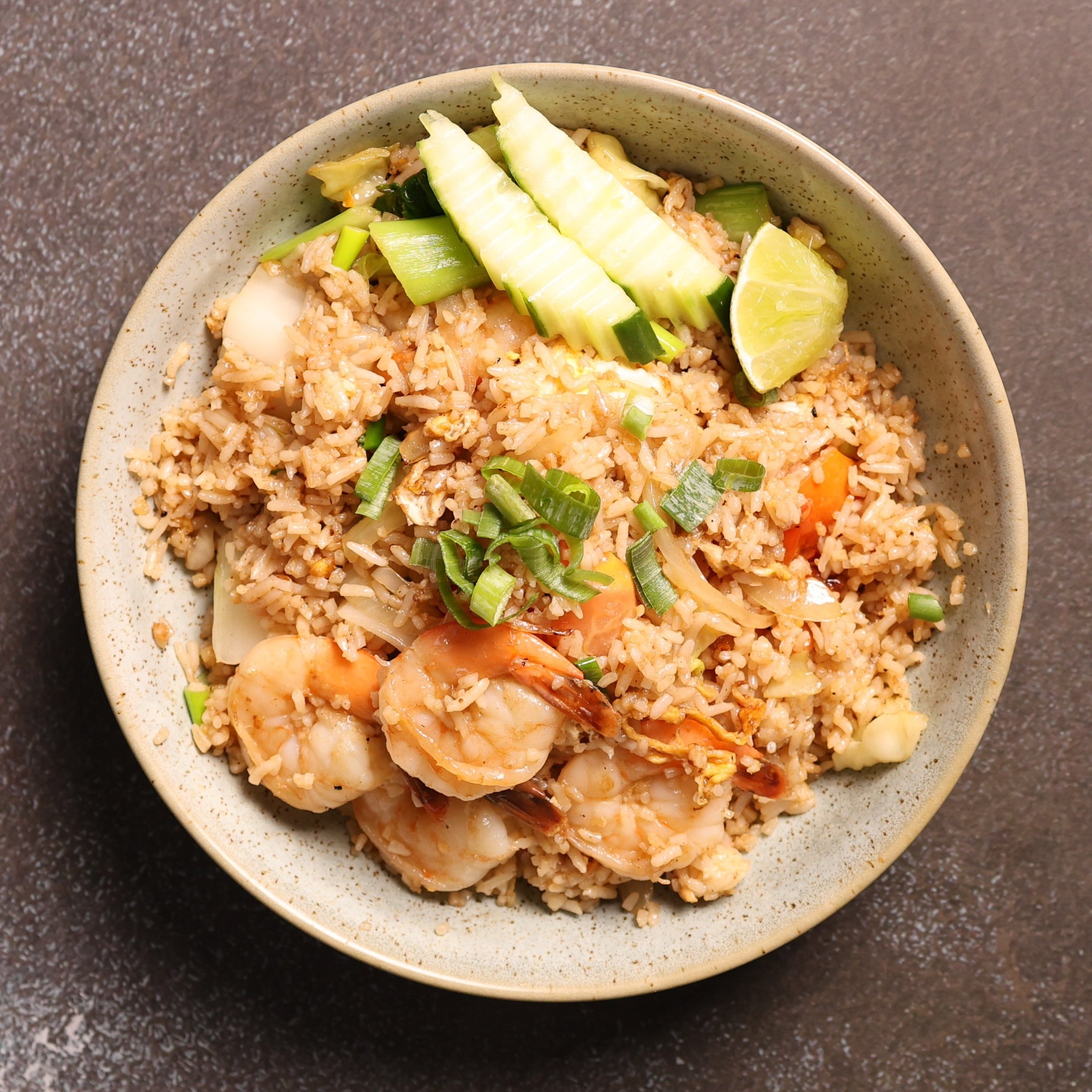 Bowl of fried rice garnished with sliced cucumber and lime on a dark countertop.