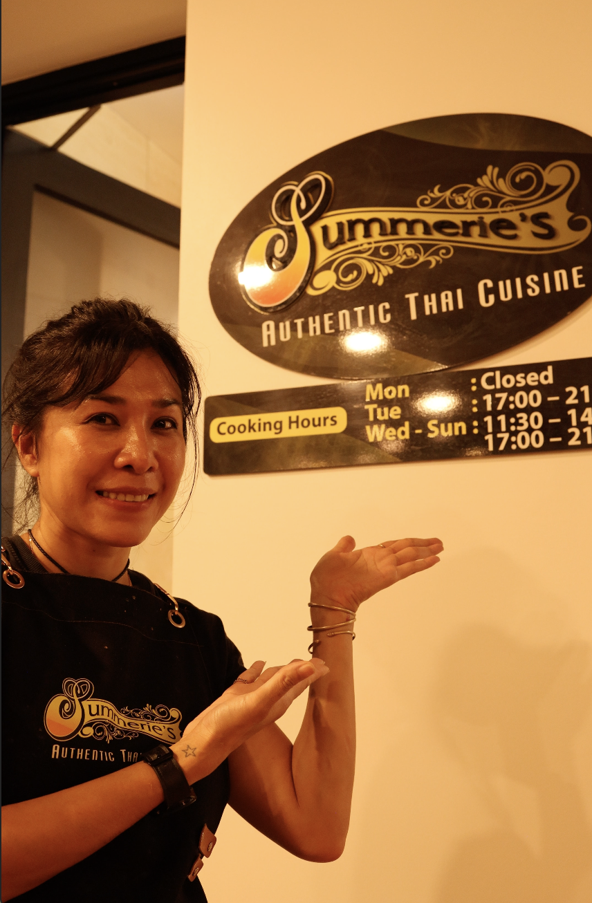 A woman smiling and pointing at a sign for Summerie's Authentic Thai Cuisine restaurant, which displays the cooking hours and closed days.