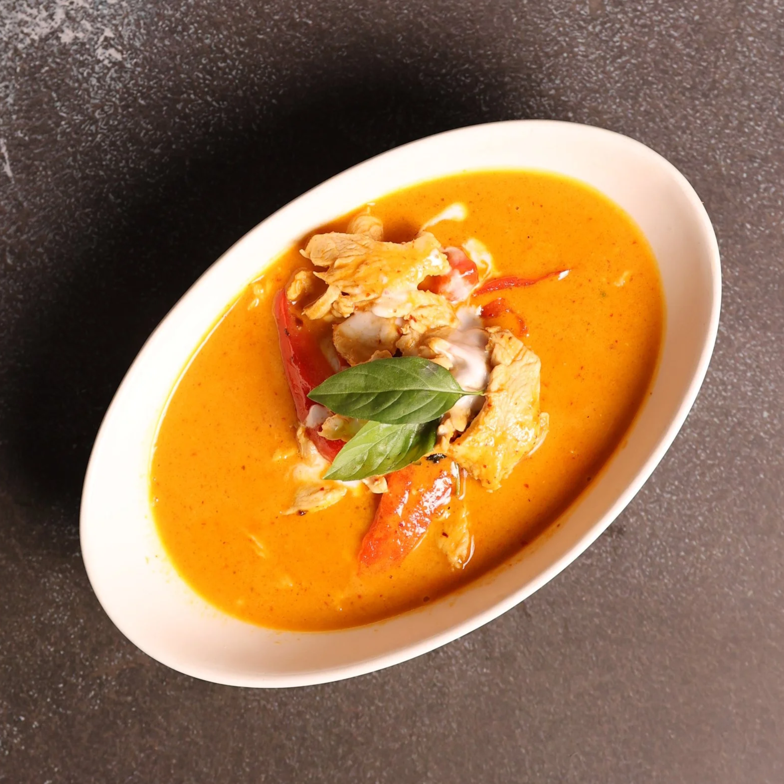 A white bowl filled with spicy red-orange curry, garnished with a fresh basil leaf, slices of red pepper, and shredded chicken. The bowl is on a dark countertop.