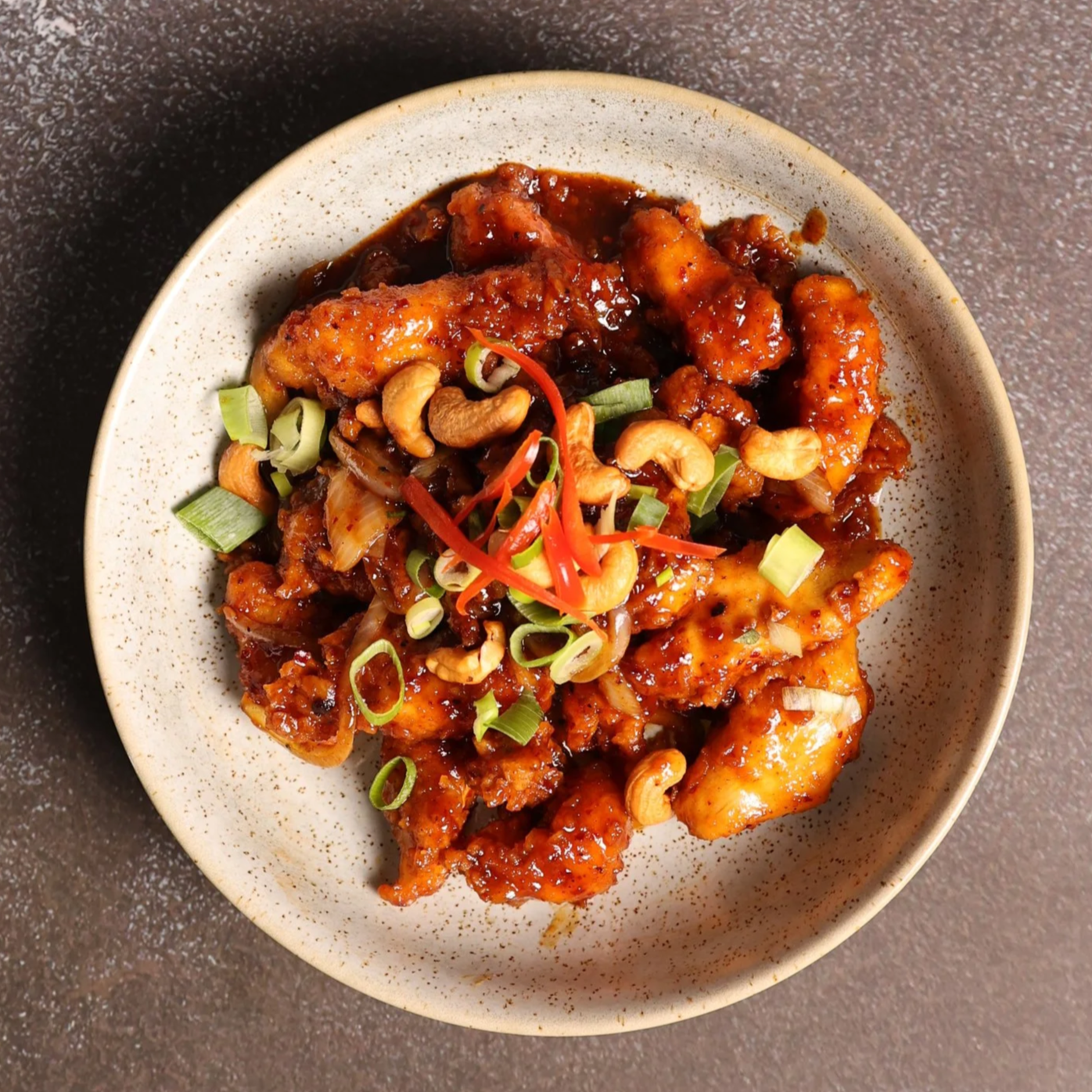 Close-up of a bowl of Asian chicken dish topped with chopped green onions, sliced red peppers, cashews, and a dark sauce.