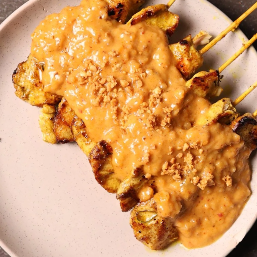 Six grilled chicken skewers topped with orange sauce and crushed peanuts served on a white plate.