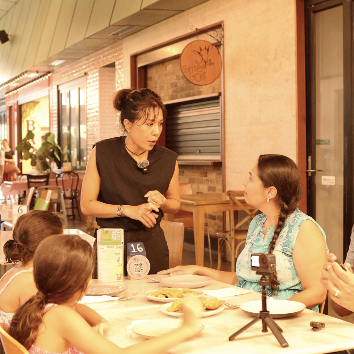 A woman in a black top is speaking to a woman in a blue dress at a restaurant table. Four young girls are seated at the table, and a small camera on a tripod is pointing at them. The restaurant has warm lighting and a rustic decor.