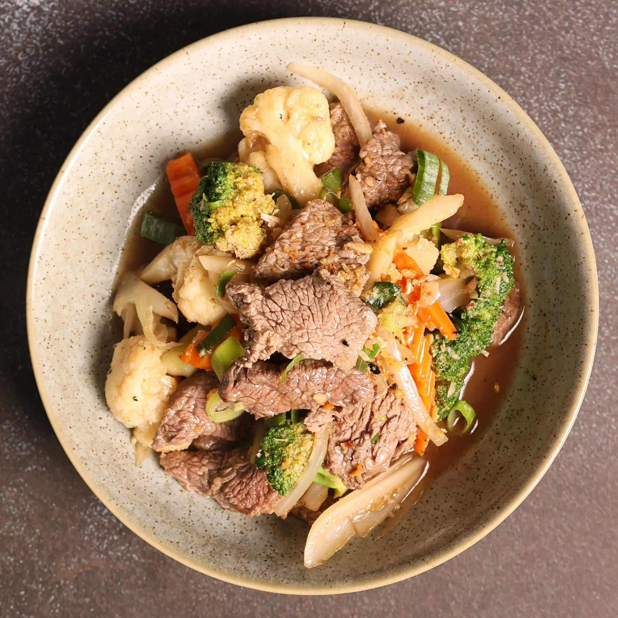A beige bowl with speckles filled with beef and vegetable soup, including broccoli, cauliflower, carrots, and green onions on a dark speckled countertop.