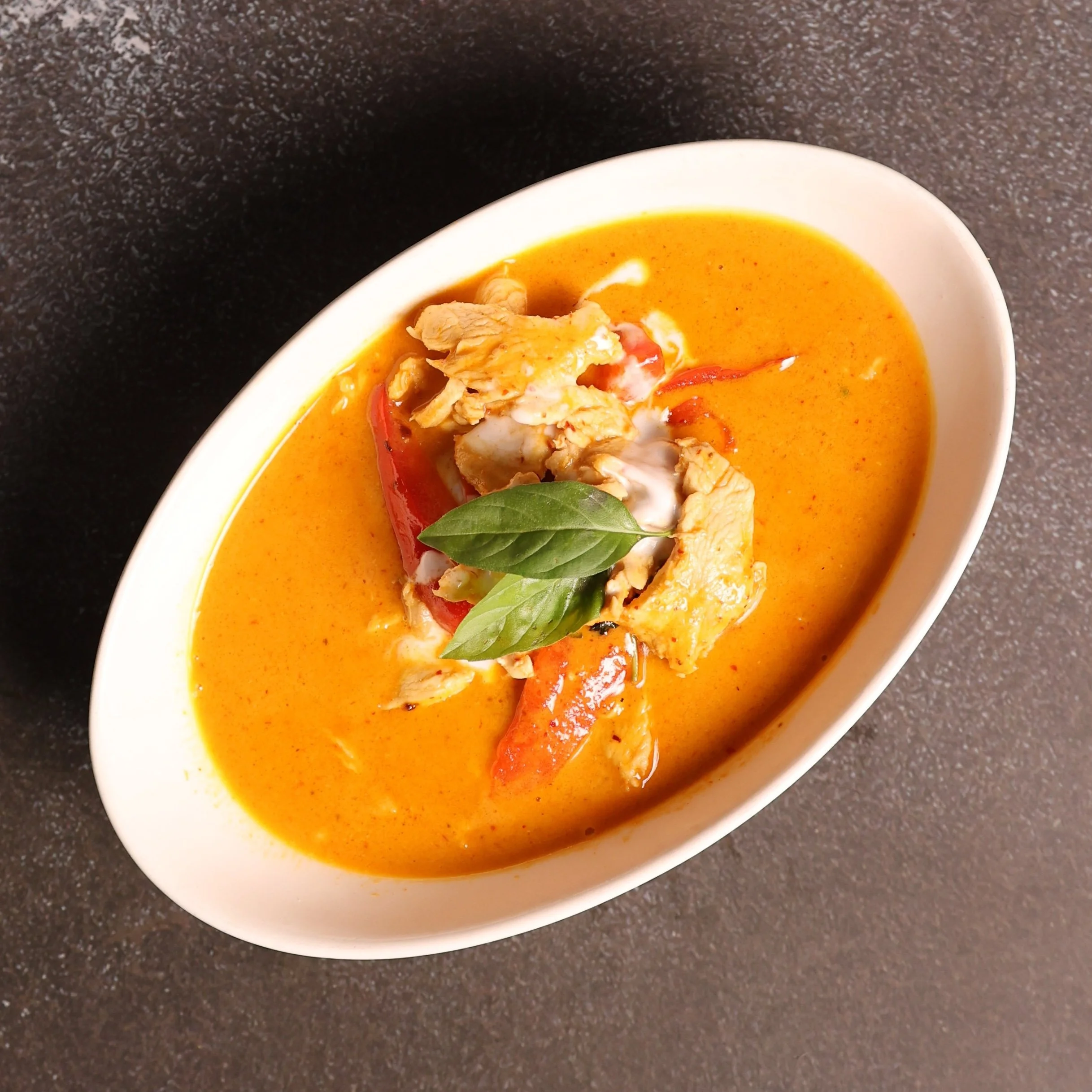 A white bowl with orange-colored soup, garnished with chopped chicken, red bell peppers, and fresh basil leaves, placed on a dark countertop.