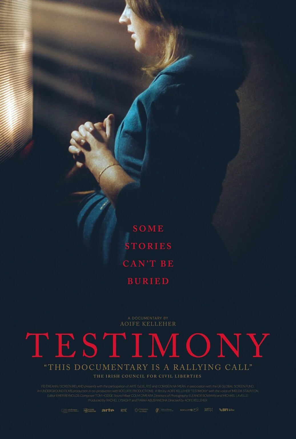 A woman with brown hair, wearing a dark blue hoodie, stands in profile with hands clasped in front of her, illuminated by a soft light. The text on the poster reads: 'Some stories can't be buried. A documentary by Aoife Kelleher. TESTIMONY. This docu