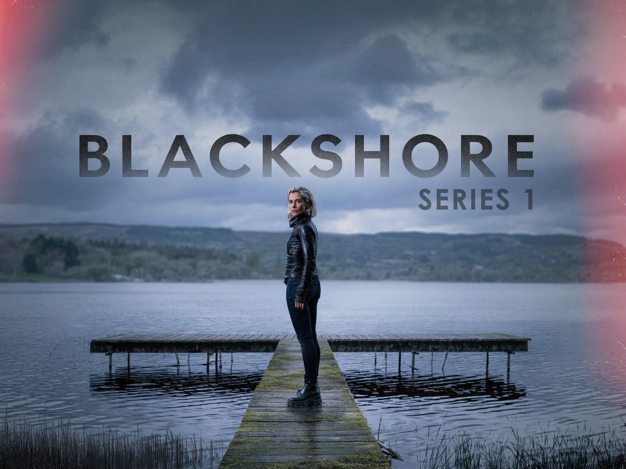 A woman standing on a wooden dock by a lake with a cloudy sky overhead and a forested landscape in the distance. The woman is wearing black leather clothing and chunky boots, looking contemplative. The image has a mysterious, moody atmosphere.