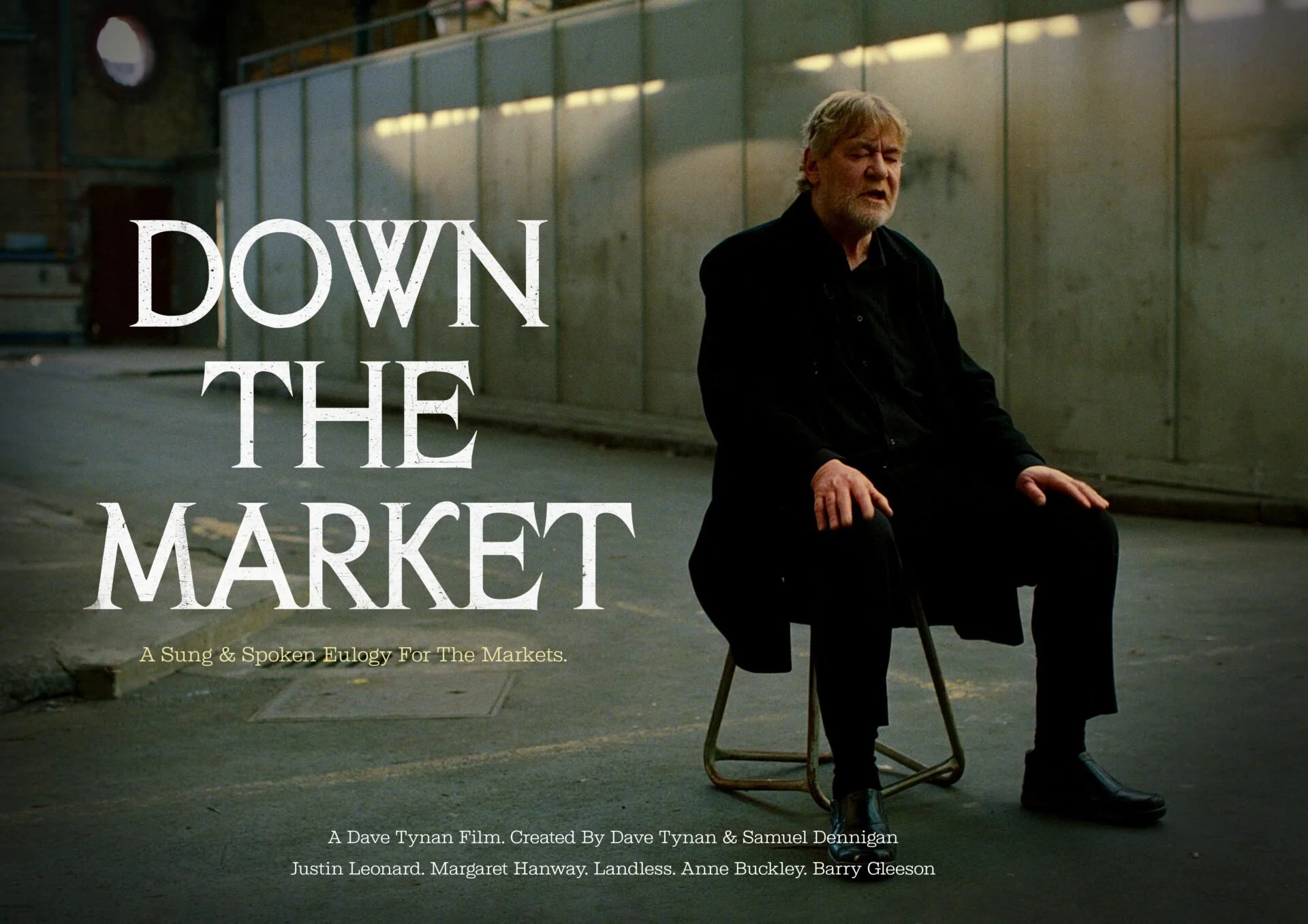 A man with a beard and gray hair wearing a black coat, sitting on a chair with his eyes closed in an industrial setting with metal walls and pipes, during the daytime.