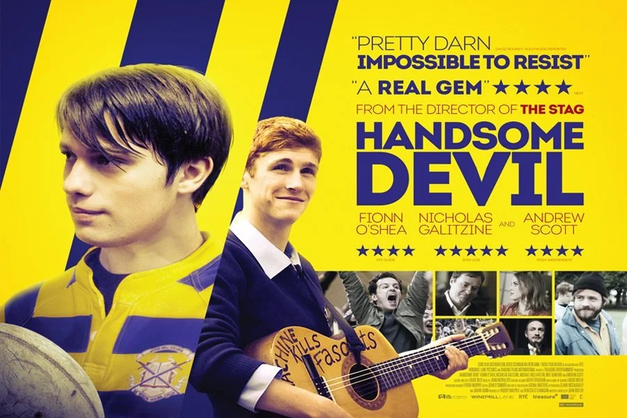 A movie poster for 'Handsome Devil' featuring two young men, one with dark hair wearing a yellow and purple jacket looking to the side, and the other with red hair wearing a blue sweater playing a guitar, against a bright yellow background with black