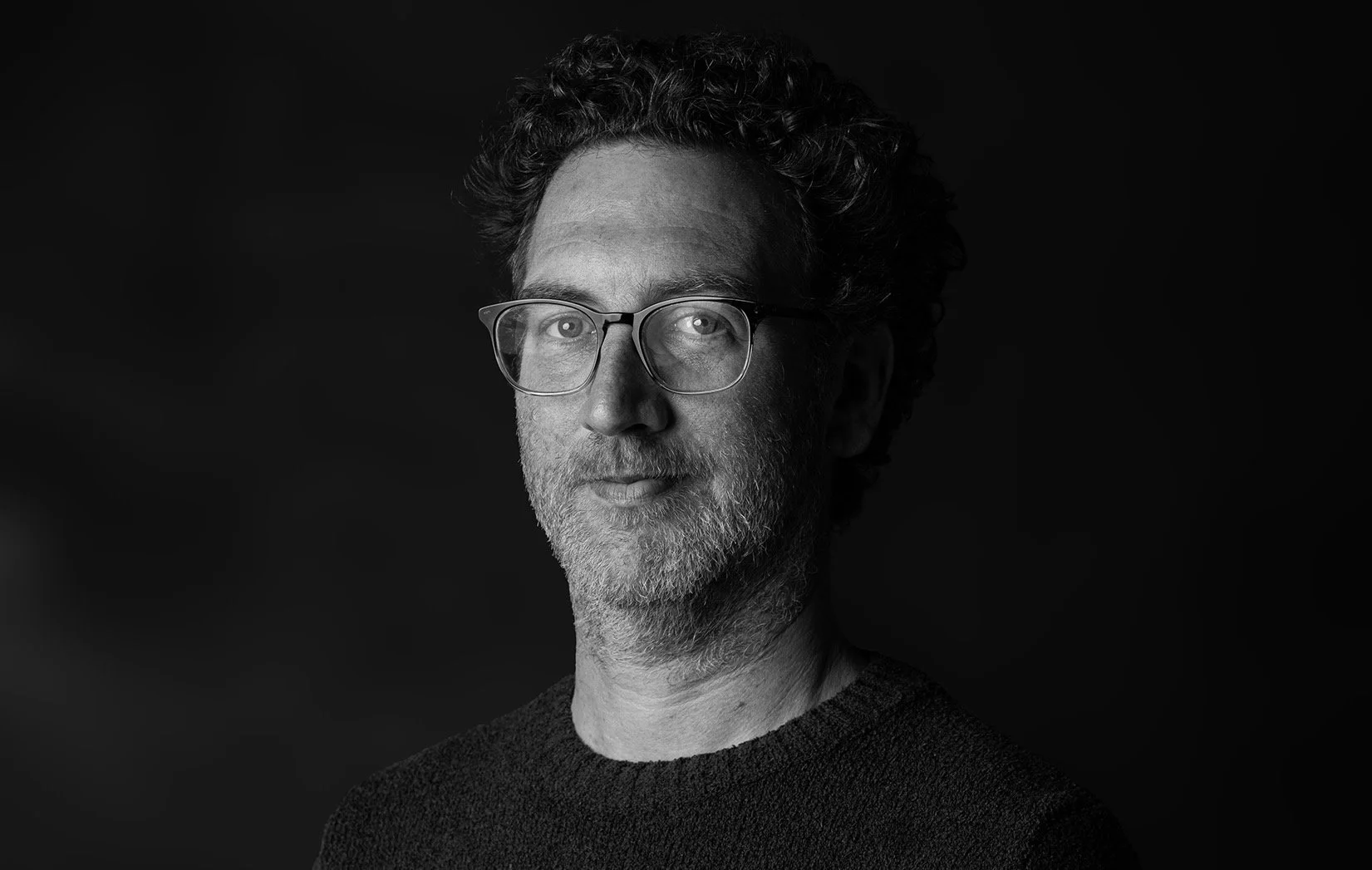 Black and white portrait of a man with curly hair, glasses, and a beard, wearing a dark sweater against a dark background.