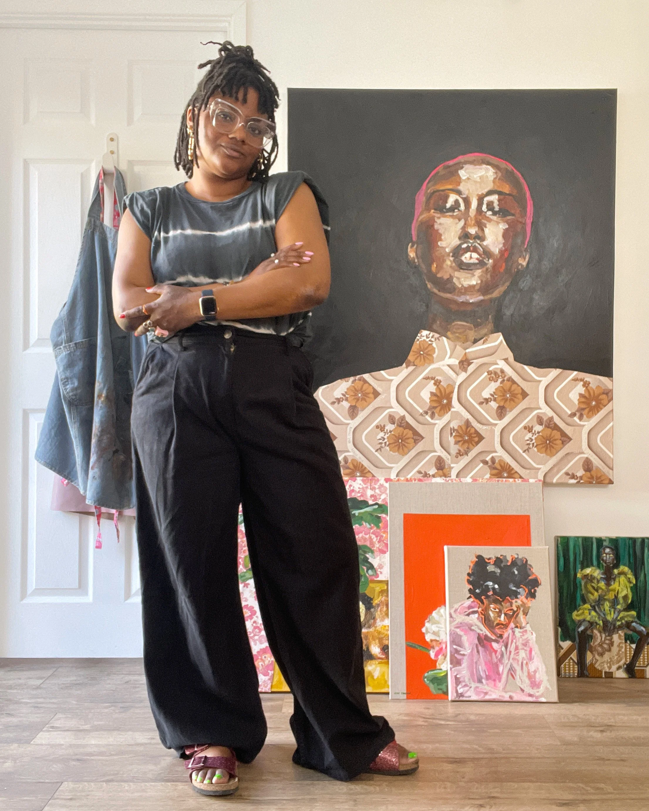 A woman standing with arms crossed in front of colorful artwork in an art gallery, wearing glasses, a tie-dye shirt, black pants, and sparkly sandals.