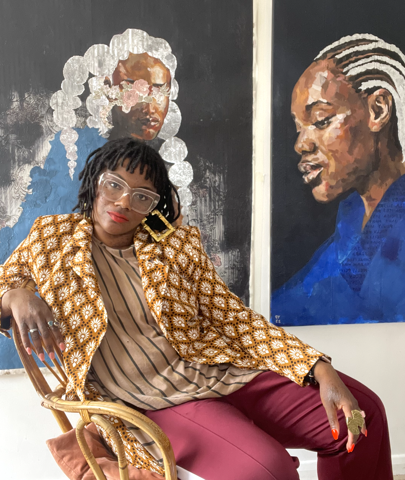 A woman with short dreadlocks, wearing glasses, a mustard yellow floral blazer, and maroon pants, sitting in front of two large portrait paintings of women with detailed hairstyles.