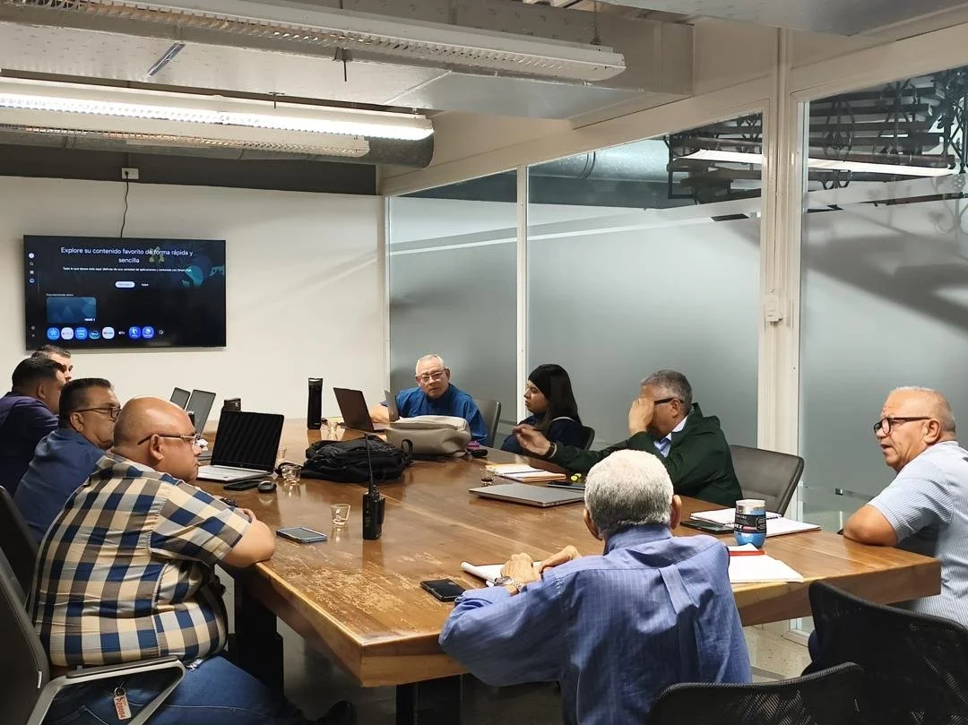 Grupo de personas en una sala de reuniones, algunos con laptops y cuadernos, participando en una discusión o presentación.
