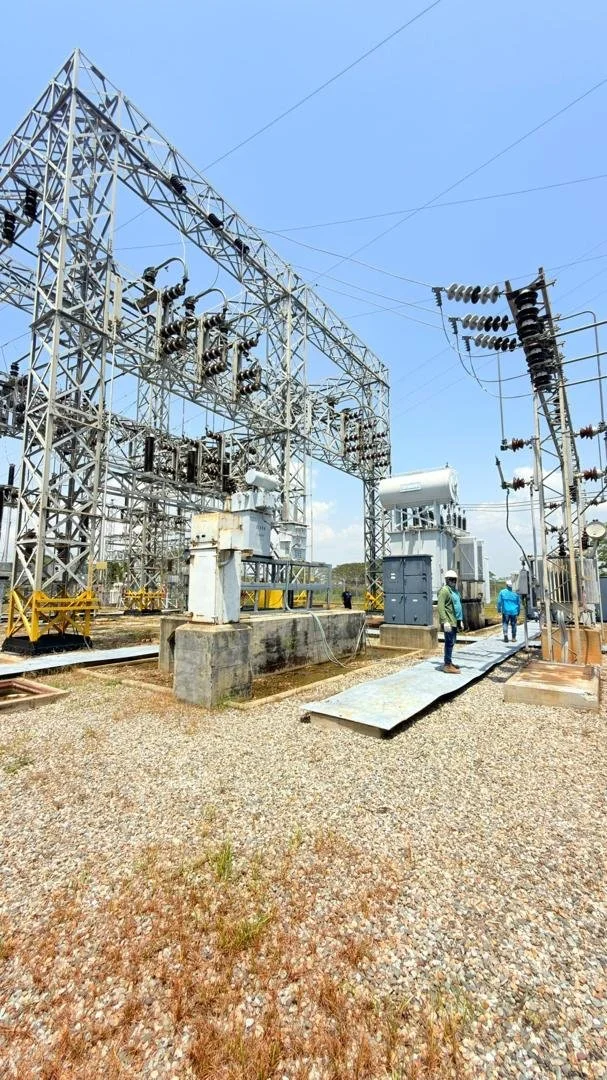 Planta de energía eléctrica con torres de transmisión y transformadores, con varias personas inspeccionando el sitio bajo un cielo despejado.