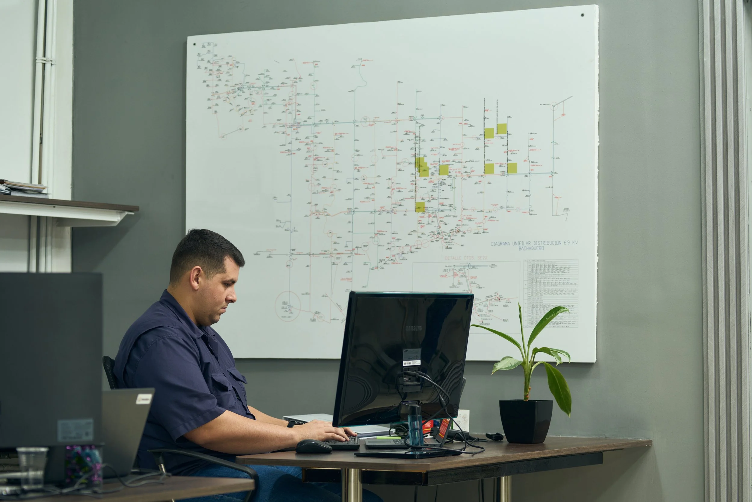 Hombre sentado en una oficina trabajando en una computadora, con un mapa técnico en la pared y una planta cerca.