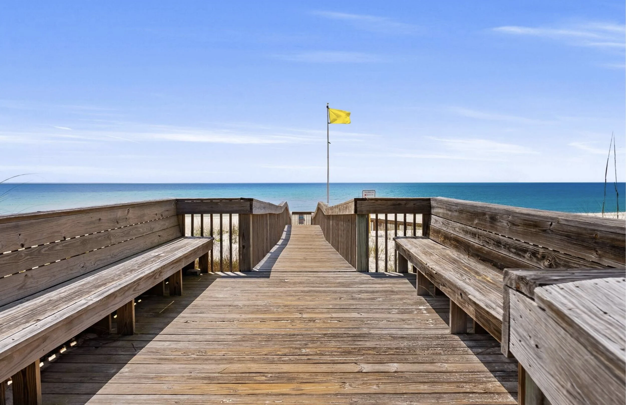 Wooden pathway with benches leading to a beach, with a yellow flag on a tall pole and blue ocean waters under a partly cloudy sky.