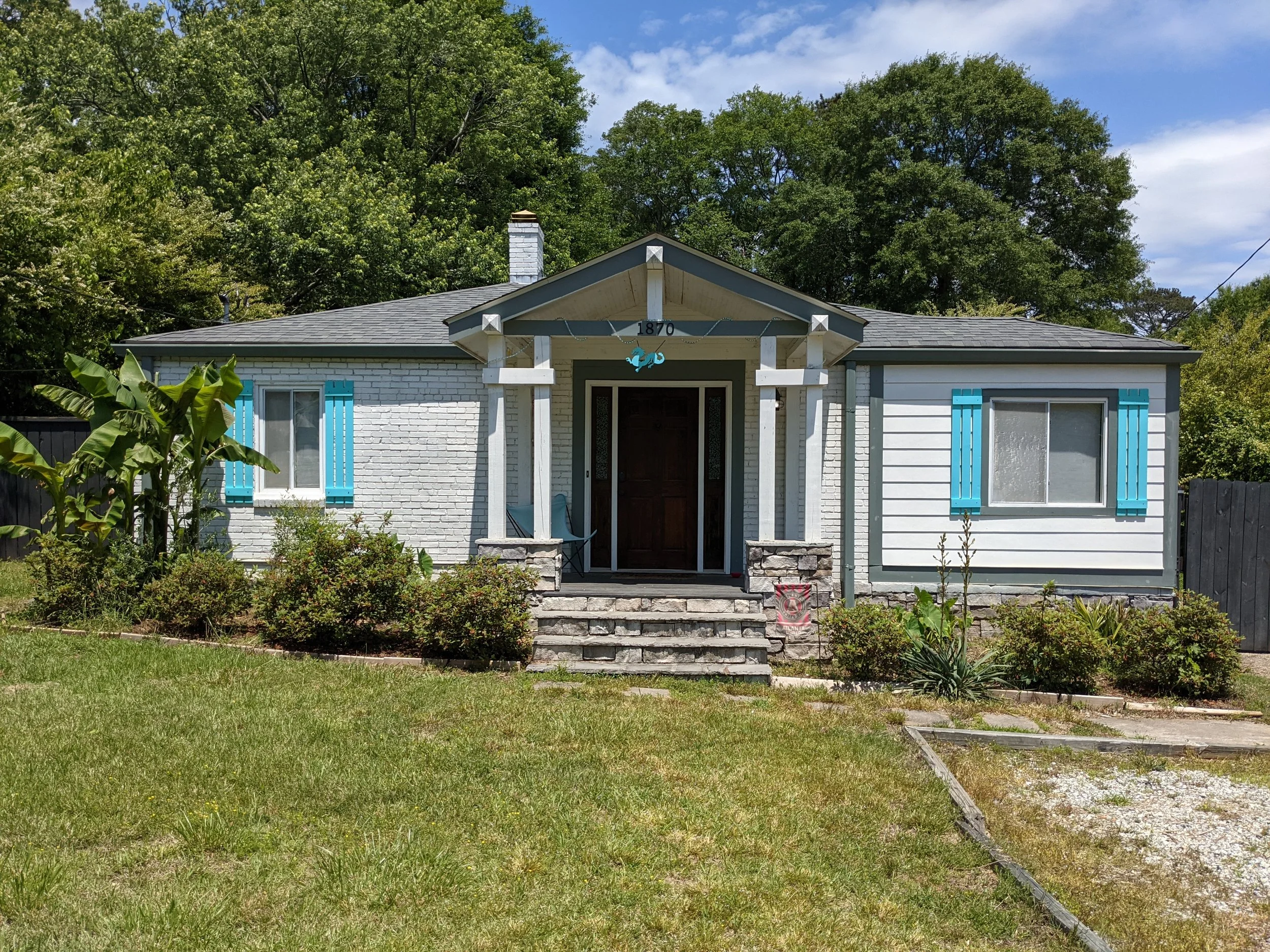 Front view of a small white brick house with blue shutters and a wooden door, surrounded by a well-maintained garden and trees in the background.