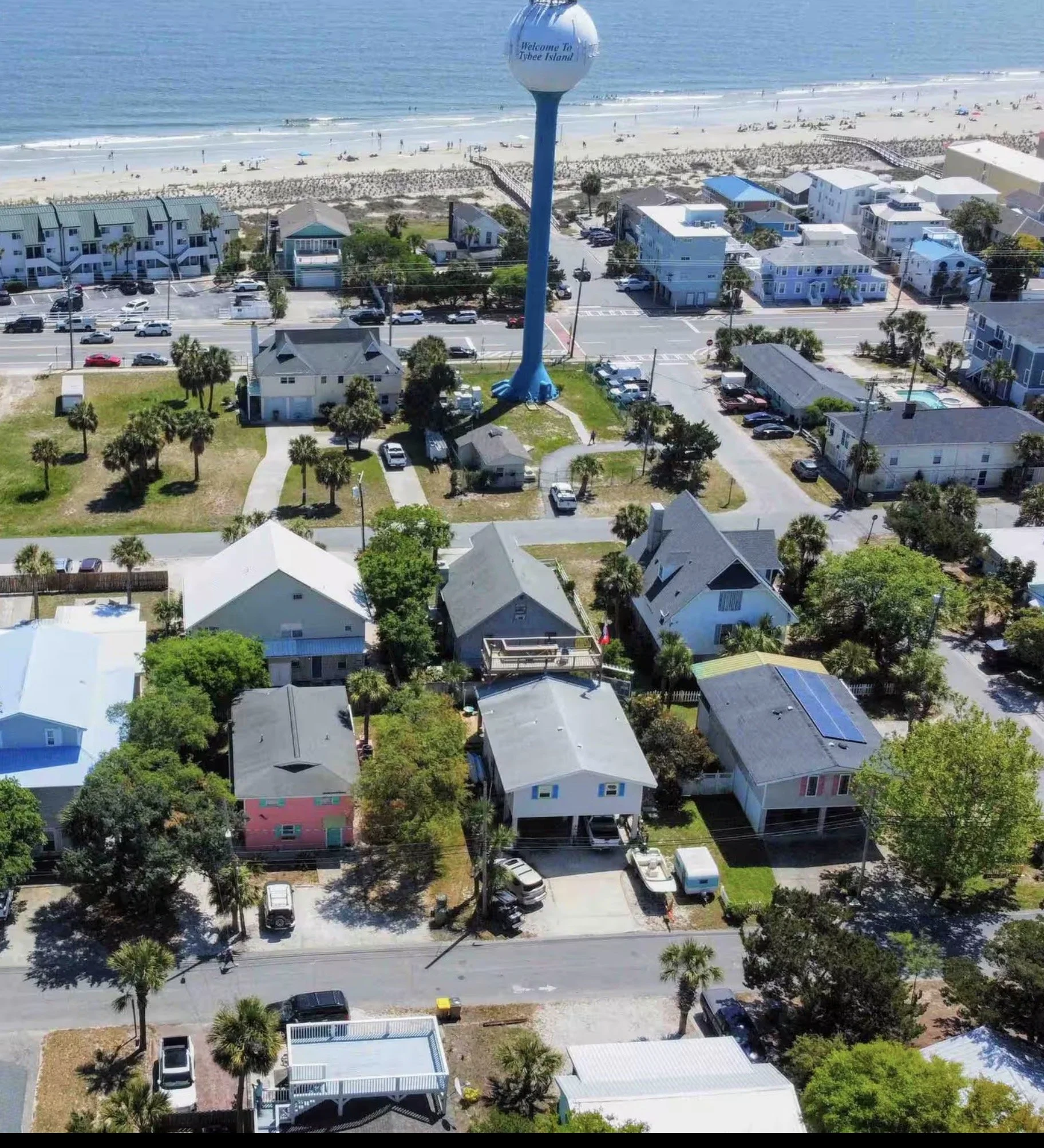 Aerial view of houses near the beach with a water tower that says 'Welcome to Tybee Island.' The ocean and sandy beach are in the background, with people walking along the shore. The residential area has trees, parked cars, and streets.