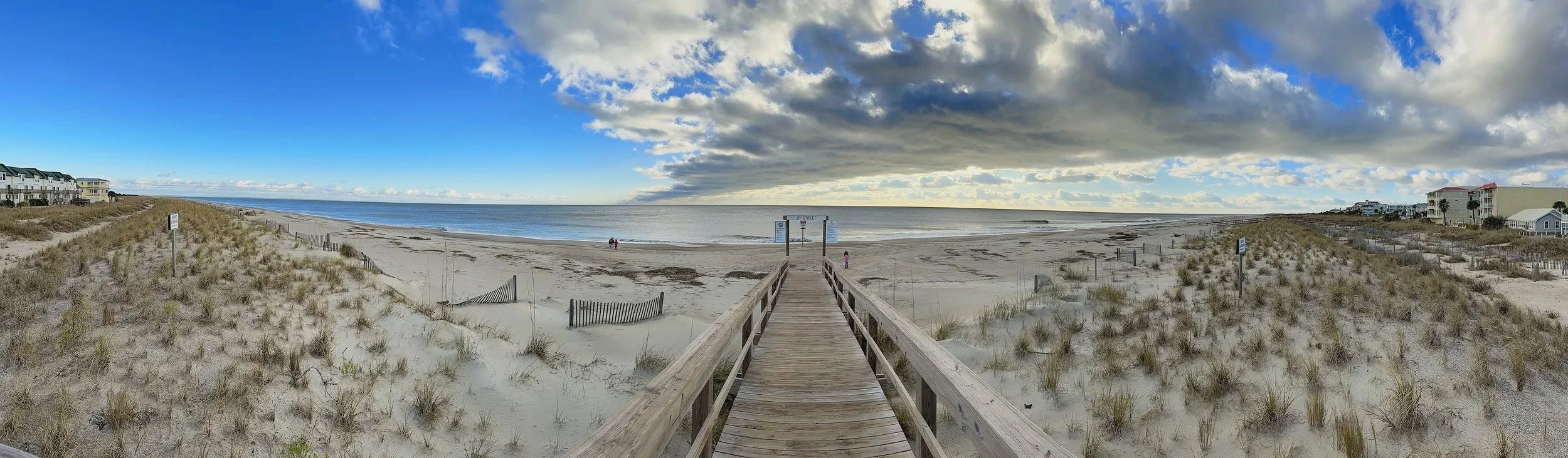 A wooden pathway leads to a sandy beach with a few people walking, bordered by beach houses on both sides, under a partly cloudy sky with sunlight breaking through clouds.