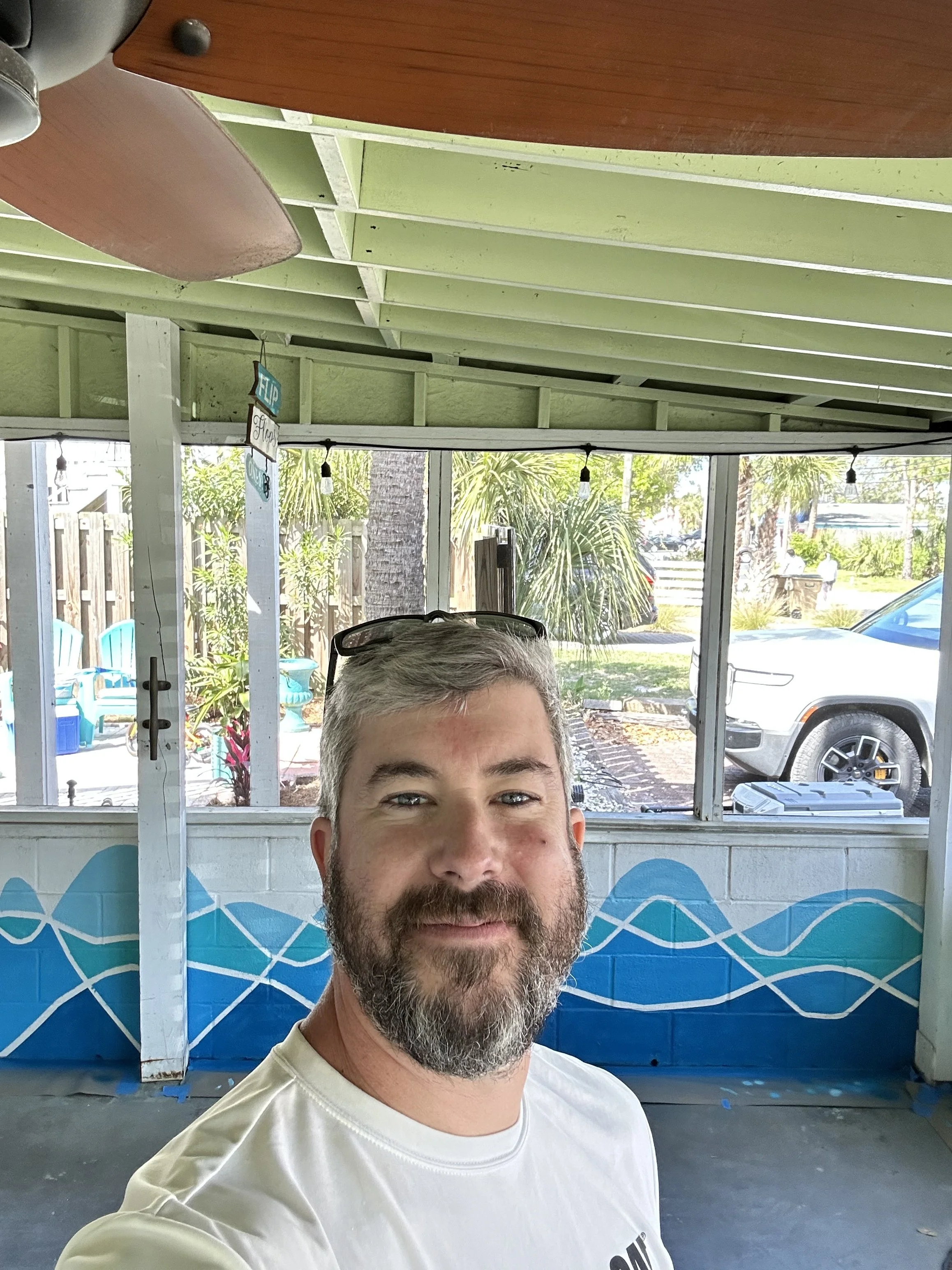 A man with gray hair and a beard smiling in front of a blue and white wave mural, with a background of outdoor plants, trees, parked cars, and a shop front.