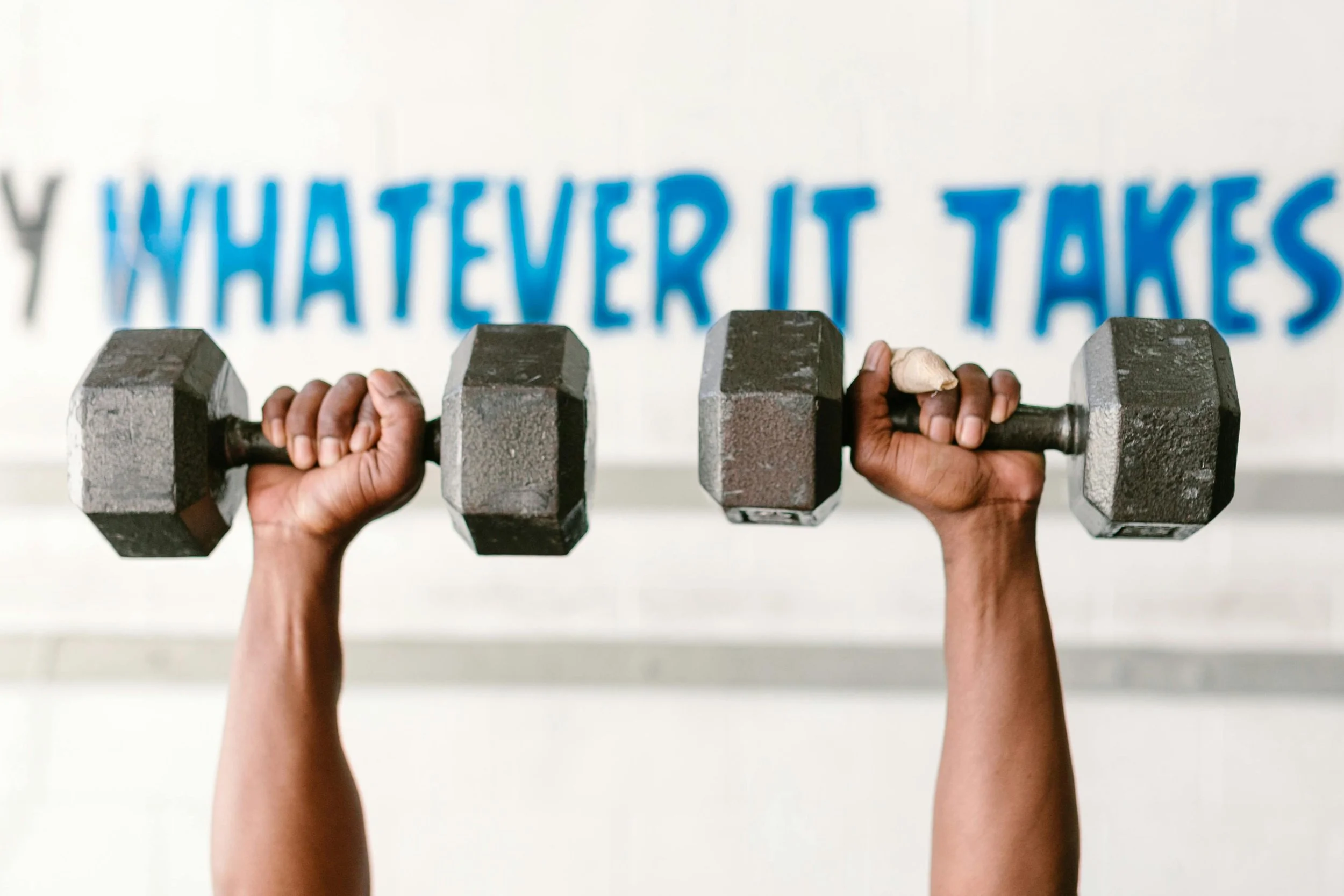 Two hands of different skin tones holding a dumbbell together, with a blurred background showing a motivational phrase on the wall.
