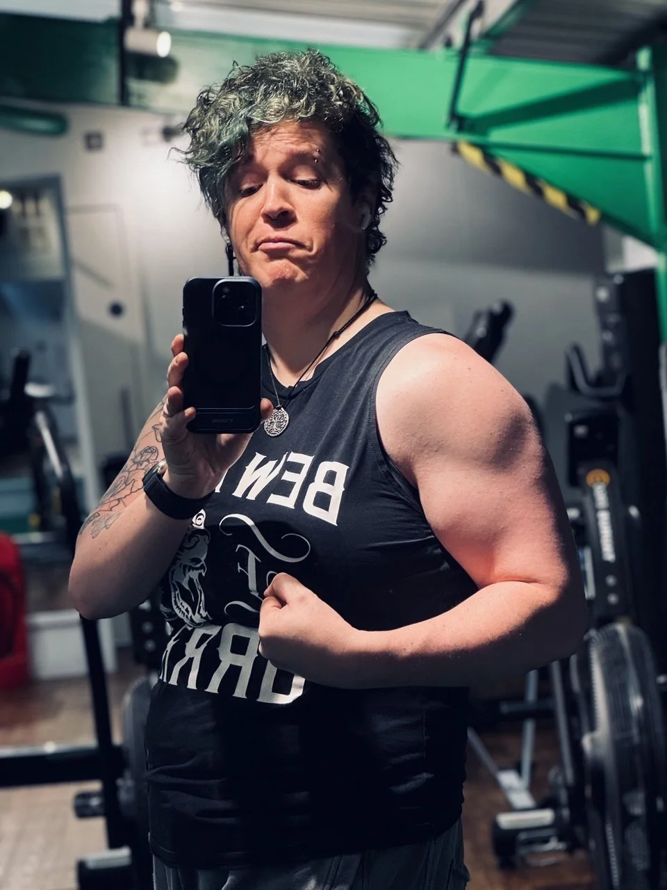 A person taking a mirror selfie in a gym. They are flexing their arm and wearing a sleeveless shirt that says 'BEW' with a graphic design on it, and have tattoos on their arm. The background shows gym equipment and a green structure.