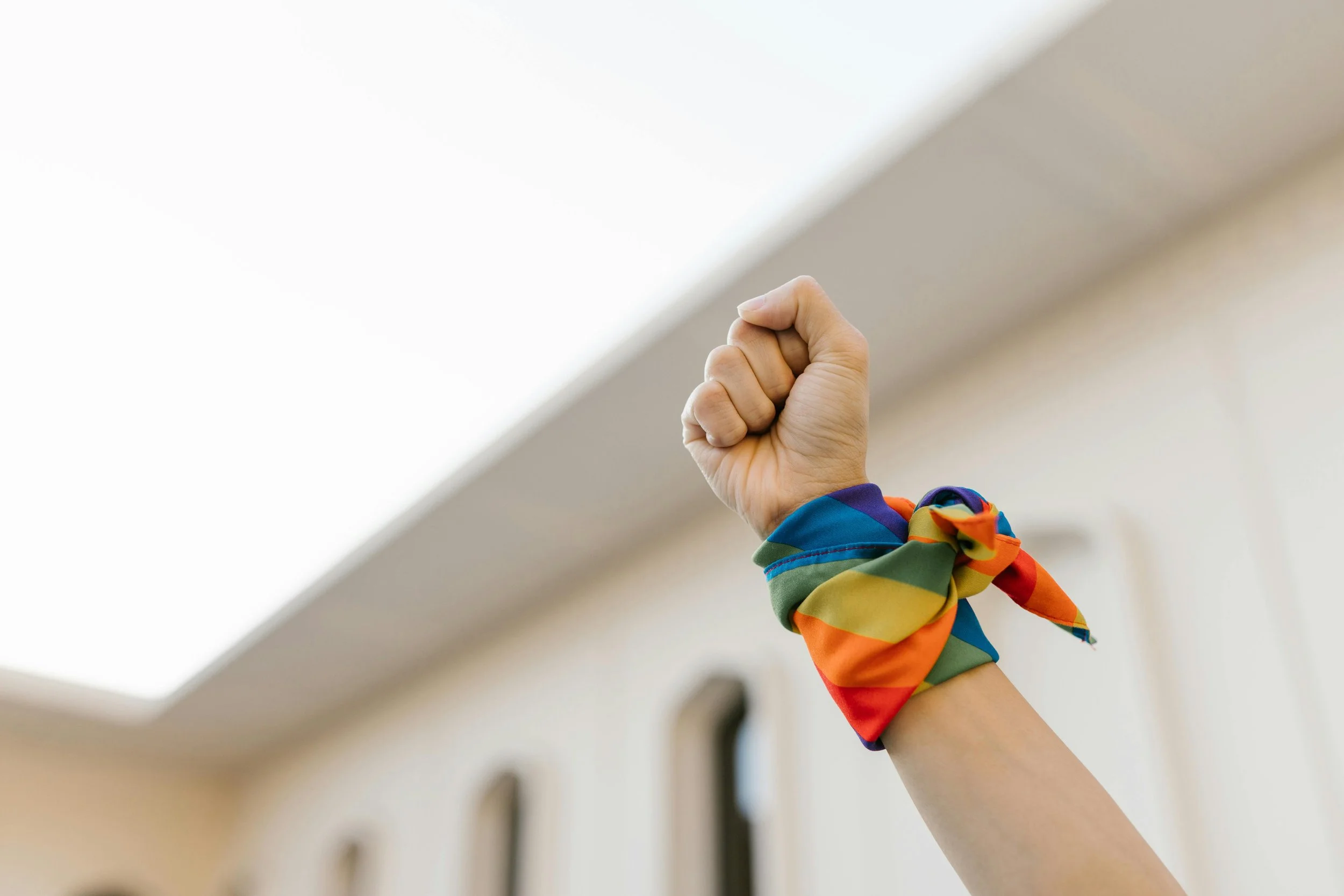 Close-up of a person's fist raised in the air, wearing a rainbow-colored rainbow ribbon and a long-sleeved shirt.