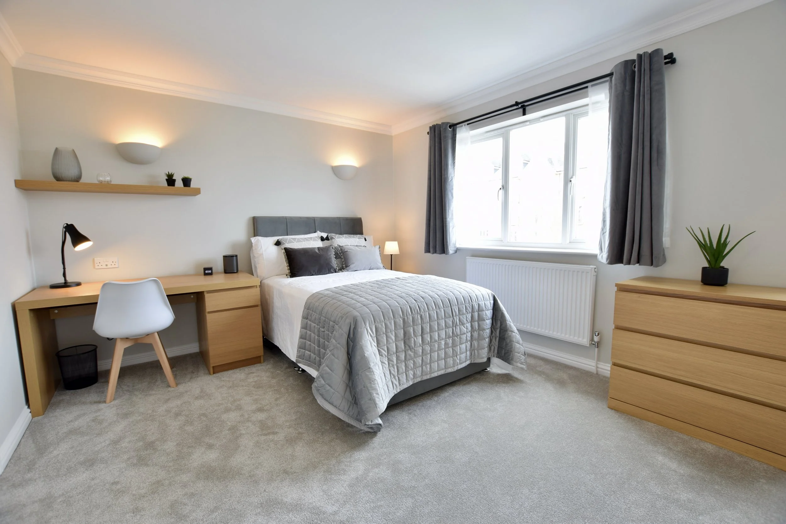 A bright bedroom with a large window and gray curtains, a bed with gray and white bedding, a wooden desk with a white chair, a black desk lamp, a small black wastebasket, and a dresser with a plant on top. The wall has two modern wall sconces.