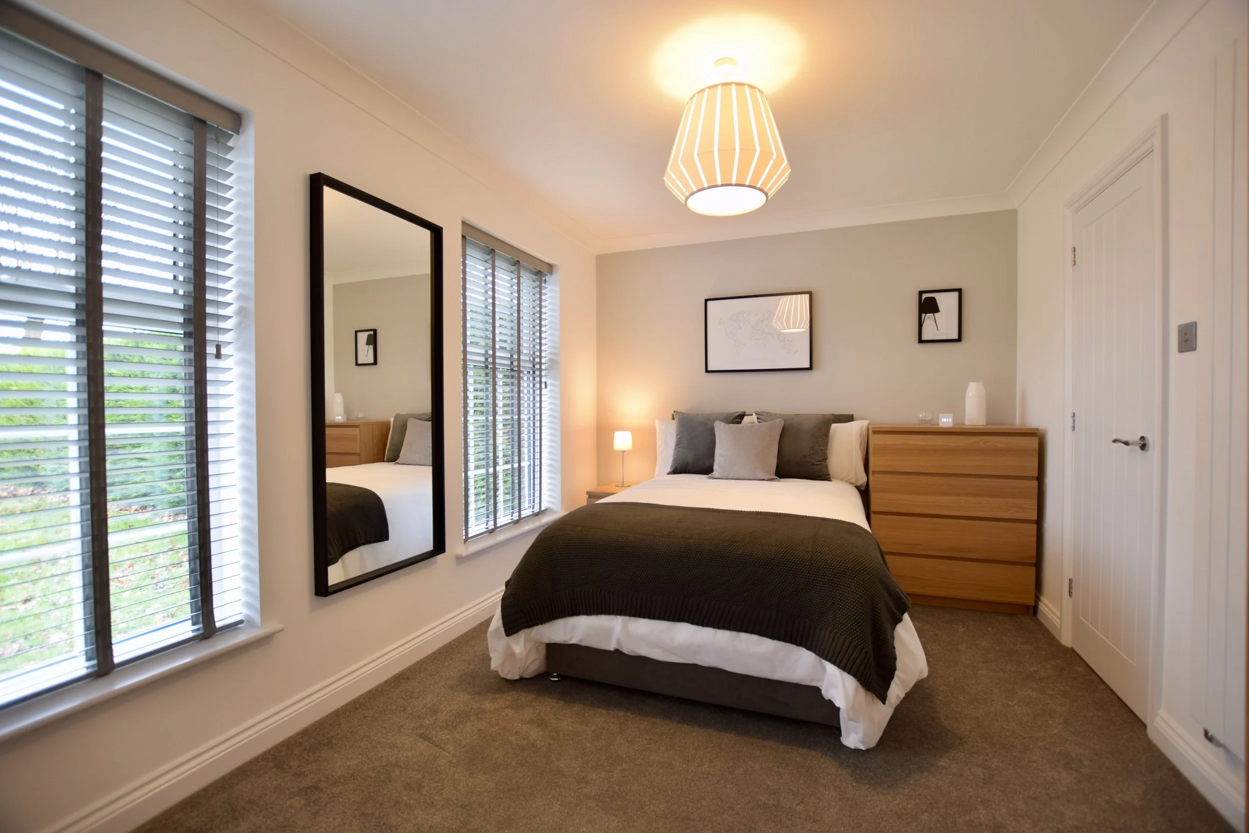 A bedroom with two large windows with white blinds, a bed with gray and white bedding, a wooden dresser, a mirror, and framed wall art. There is a bedside lamp and soft lighting from a ceiling fixture.