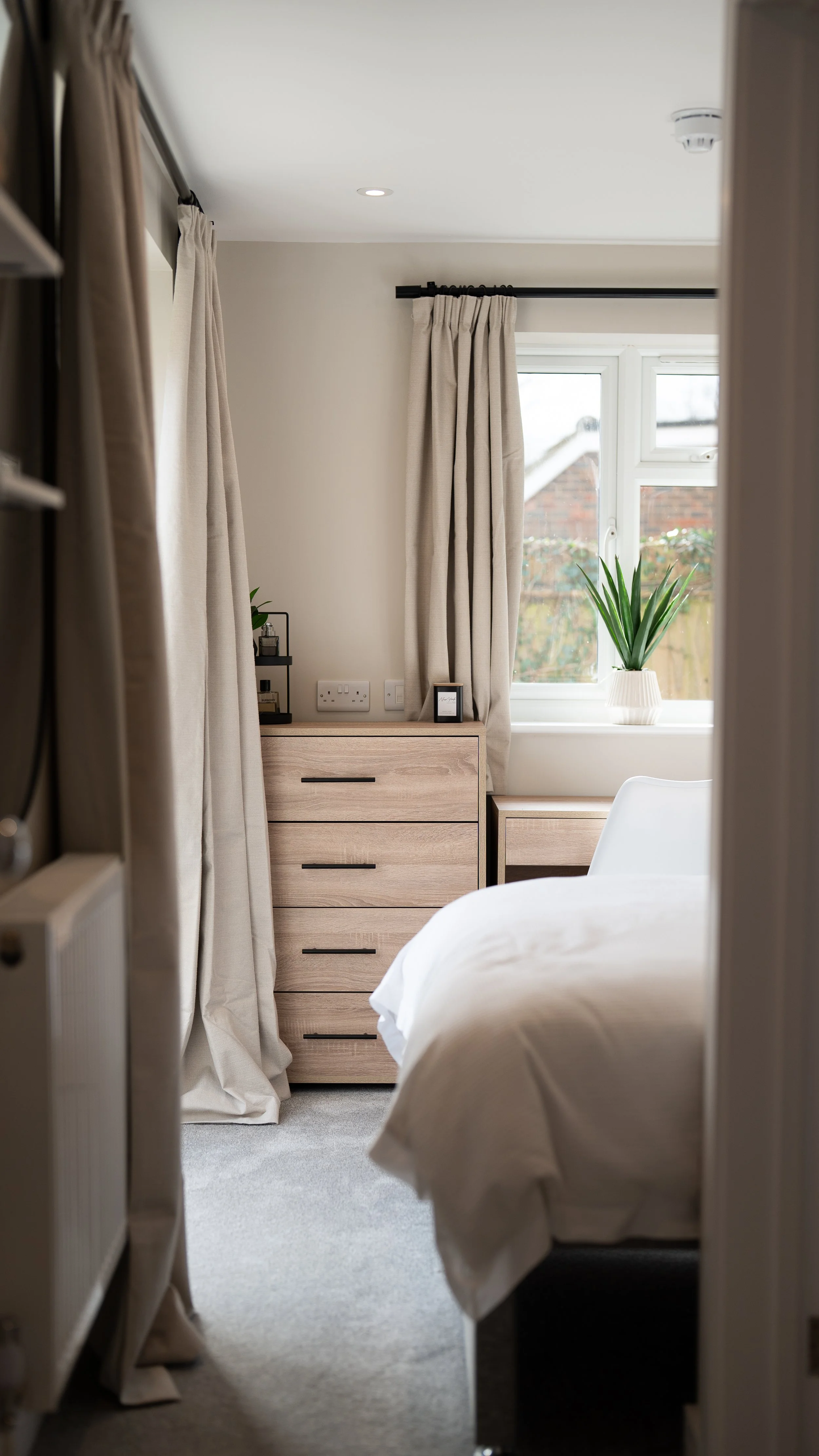 View of a bright bedroom with beige curtains, a window with a potted plant, and a light wood dresser.