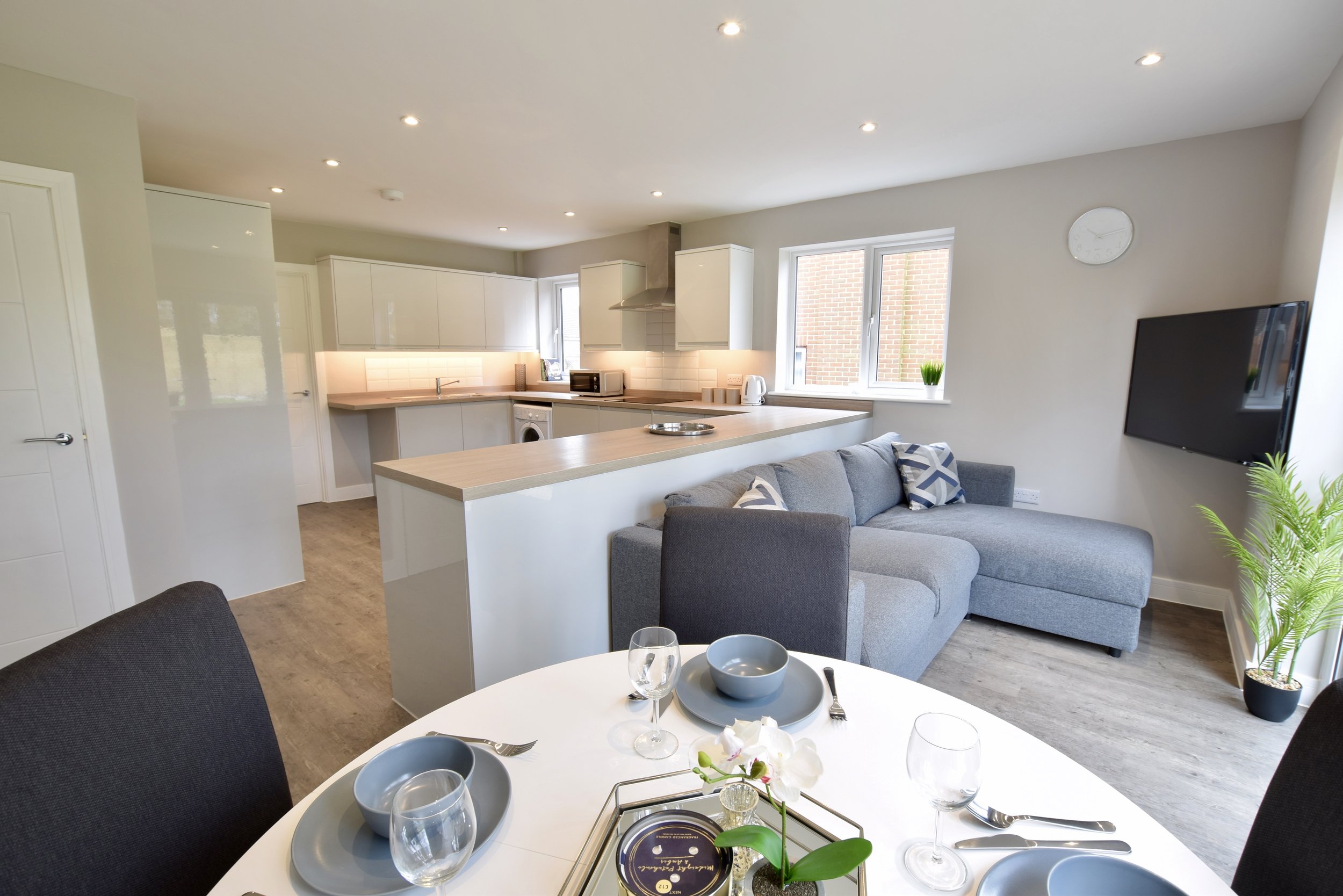 Open-concept kitchen and living room with modern decor, white cabinets, a gray sofa, a dining table set for breakfast, large windows, and a flat-screen TV on the wall.