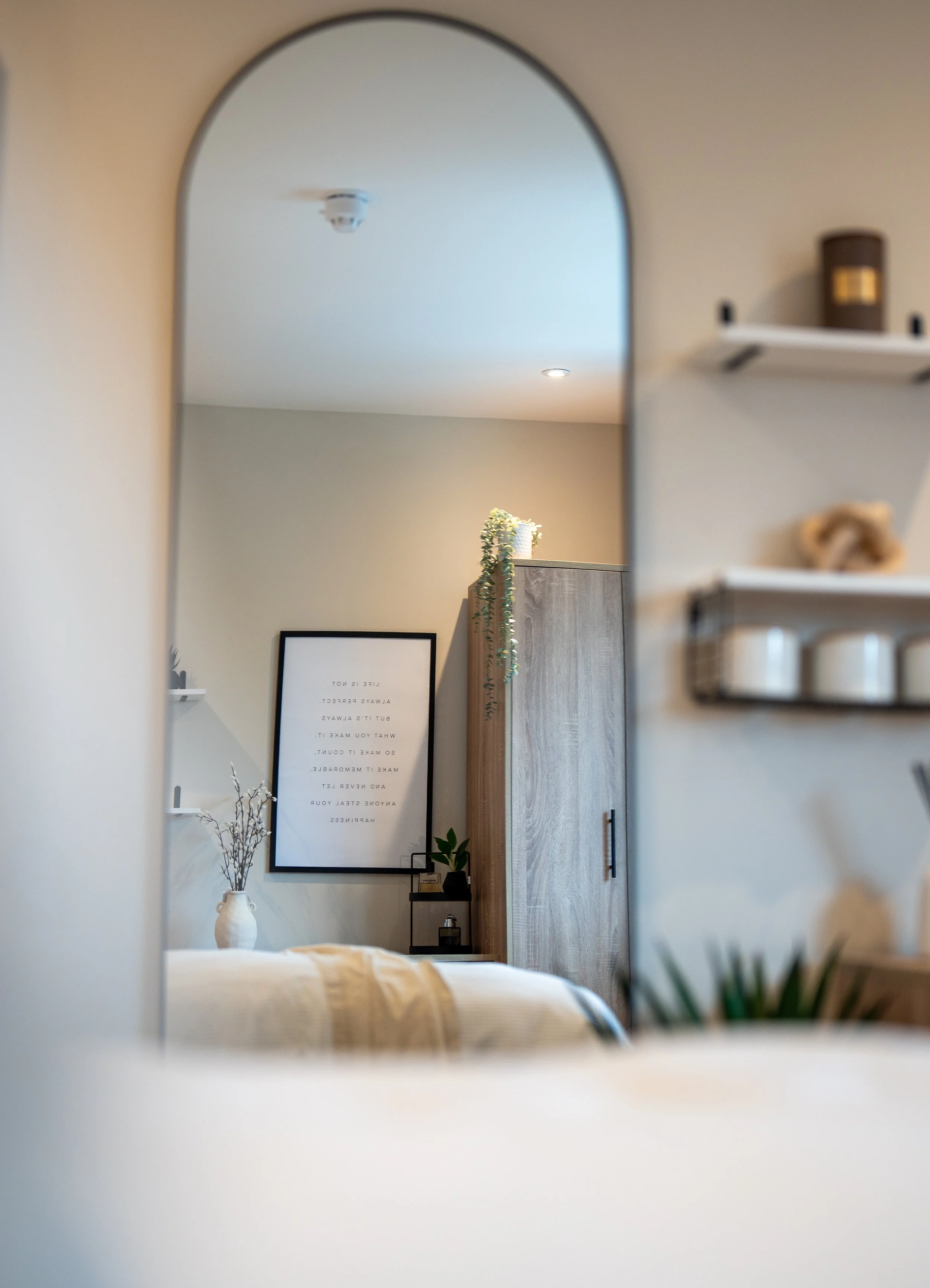 A bedroom view through a mirror, showing a bed with beige bedding, a white vase with branches, a framed quote, a wooden cabinet with a green plant, and wall shelves with decorative items.