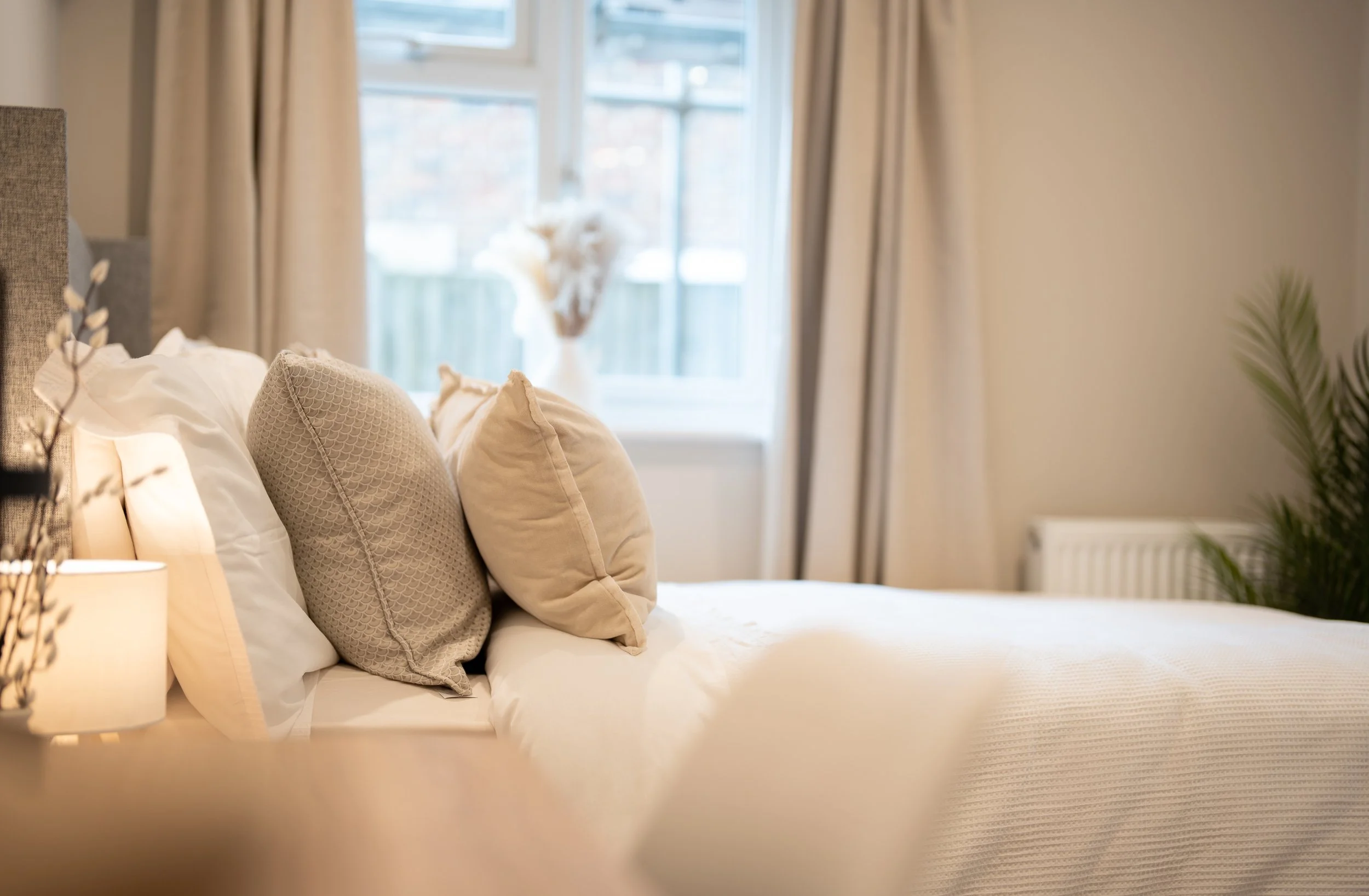 A neatly made bed with beige pillows and a cream bedspread in a bedroom with large windows and light-colored curtains.