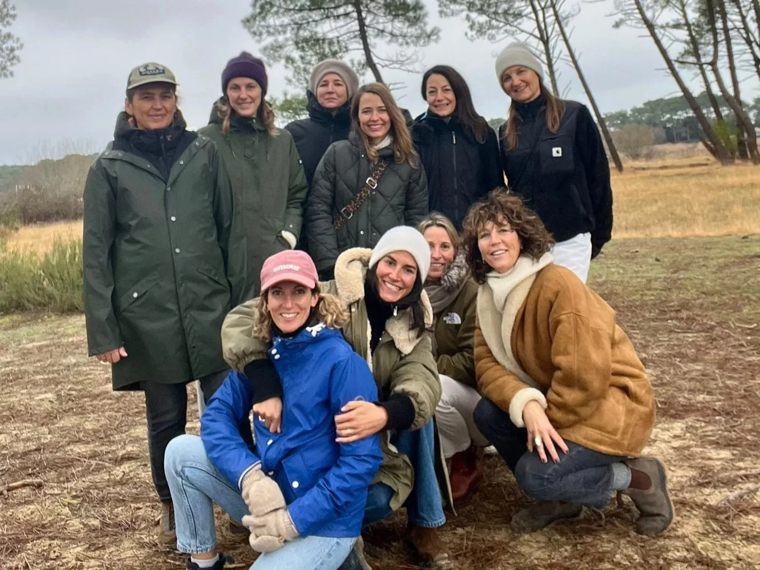 Groupe de neuf femmes posant dans la nature, par temps humide, portant des vêtements chauds et des accessoires d'extérieur.