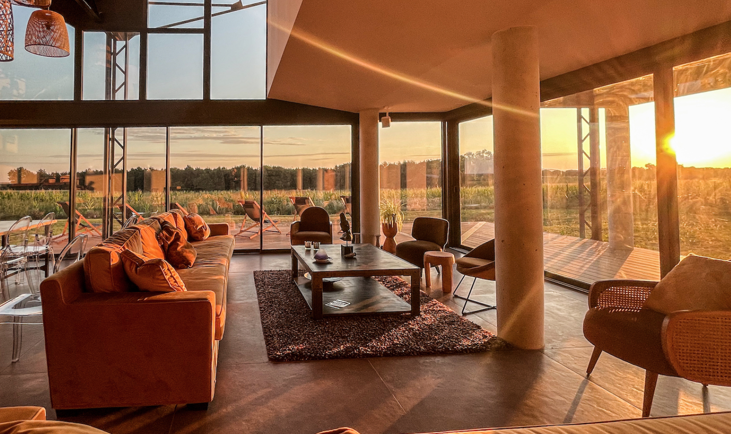Salon avec grandes fenêtres donnant sur un paysage rural au coucher du soleil, mobilier en bois, canapés et chaises confortables, tapis tissé, décor moderne et chaleureux.