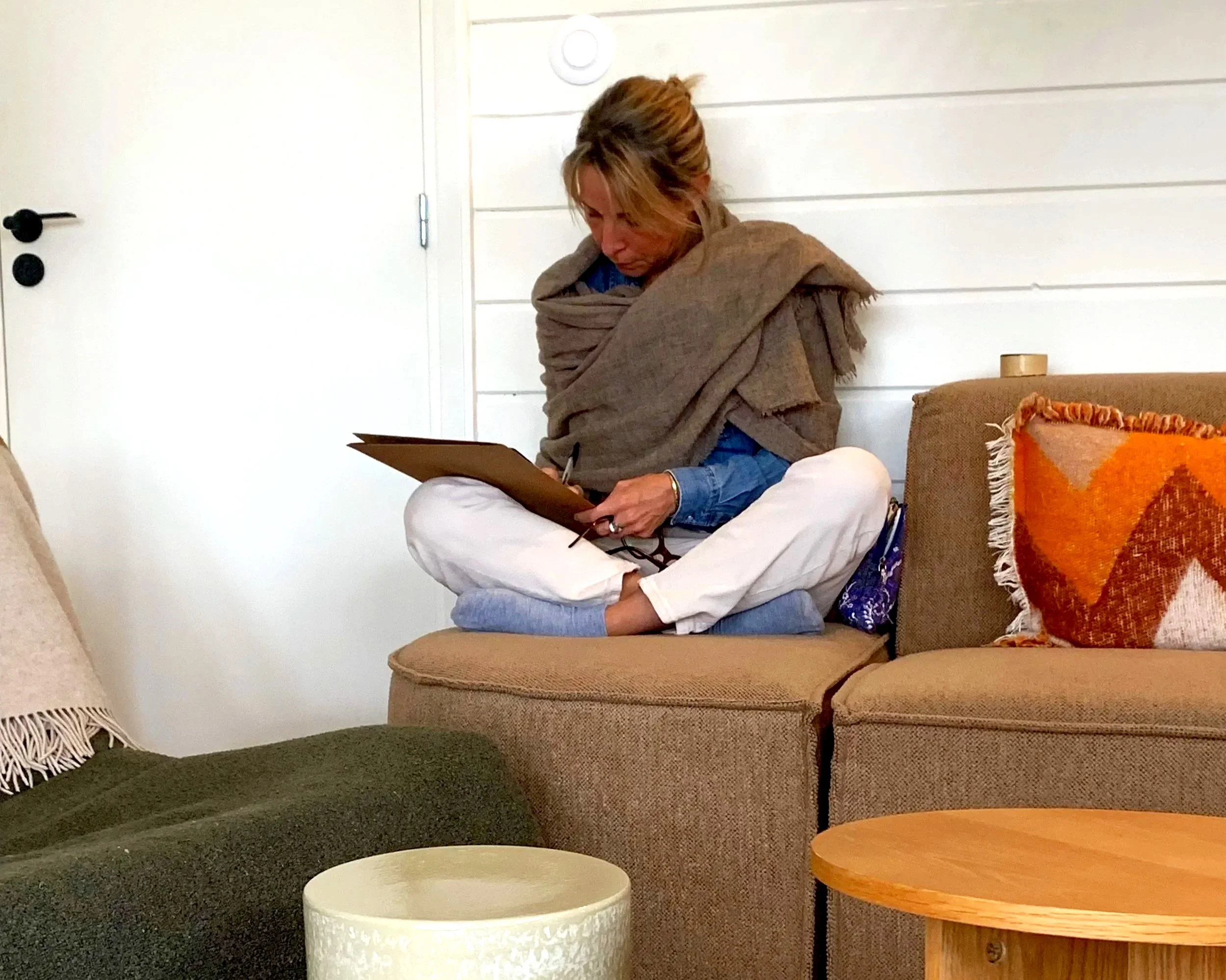 Une femme assise sur un canapé beige, portant une shawl marron, lisant ou écrivant sur un carnet. La pièce a des murs blancs avec un décor minimaliste, un coussin orange et un mur blanc en arrière-plan.
