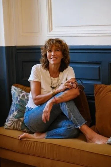 Une femme assise sur un canapé, portant un t-shirt blanc, un jean et tenant ses lunettes, dans un intérieur moderne avec des murs beige et bleu.