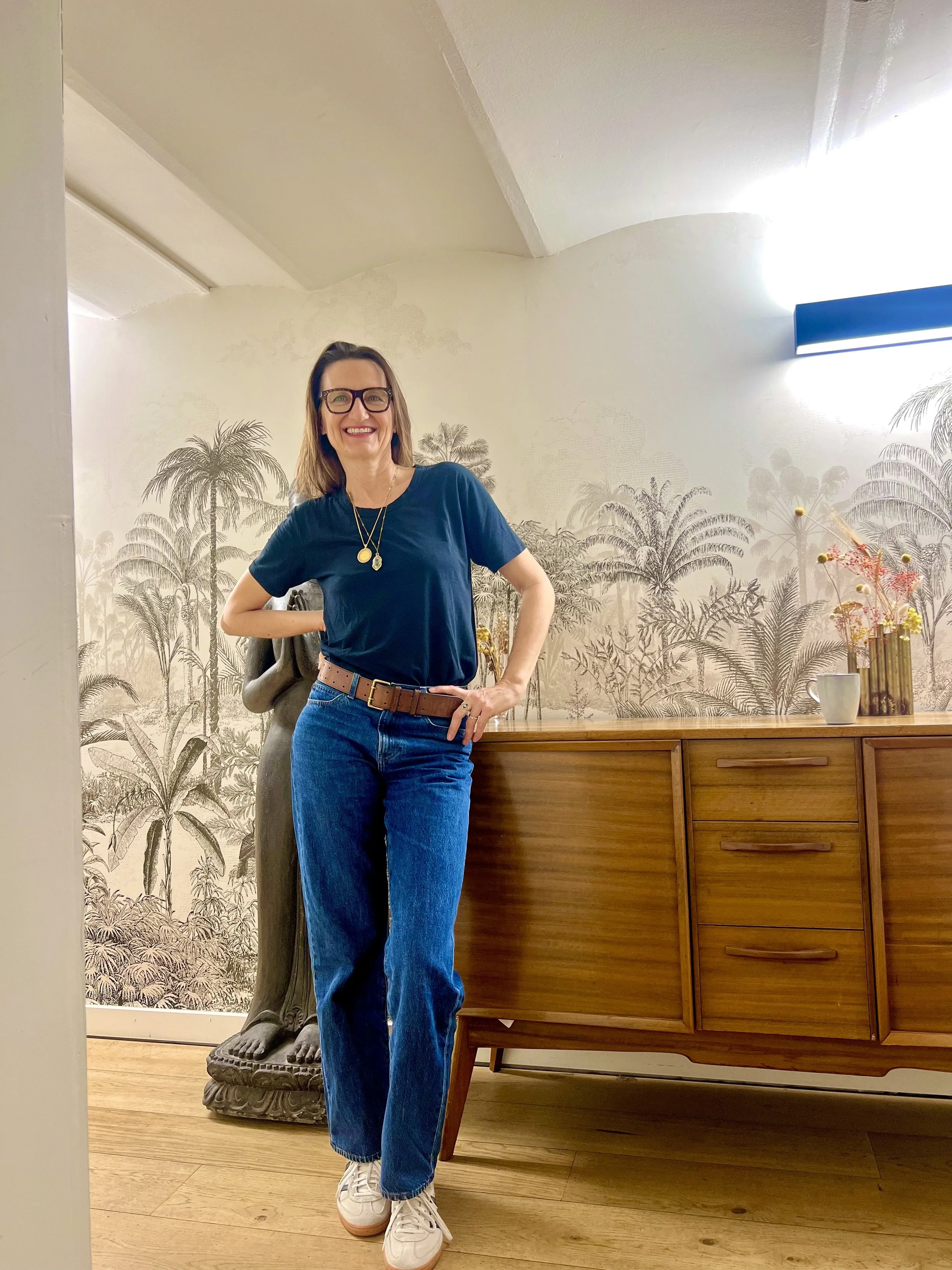 Femme souriante portant une chemise bleue, des lunettes, un collier, des jeans et des baskets blanches, posant dans une maison avec un mur décoré de palmier en arrière-plan et un meuble en bois.