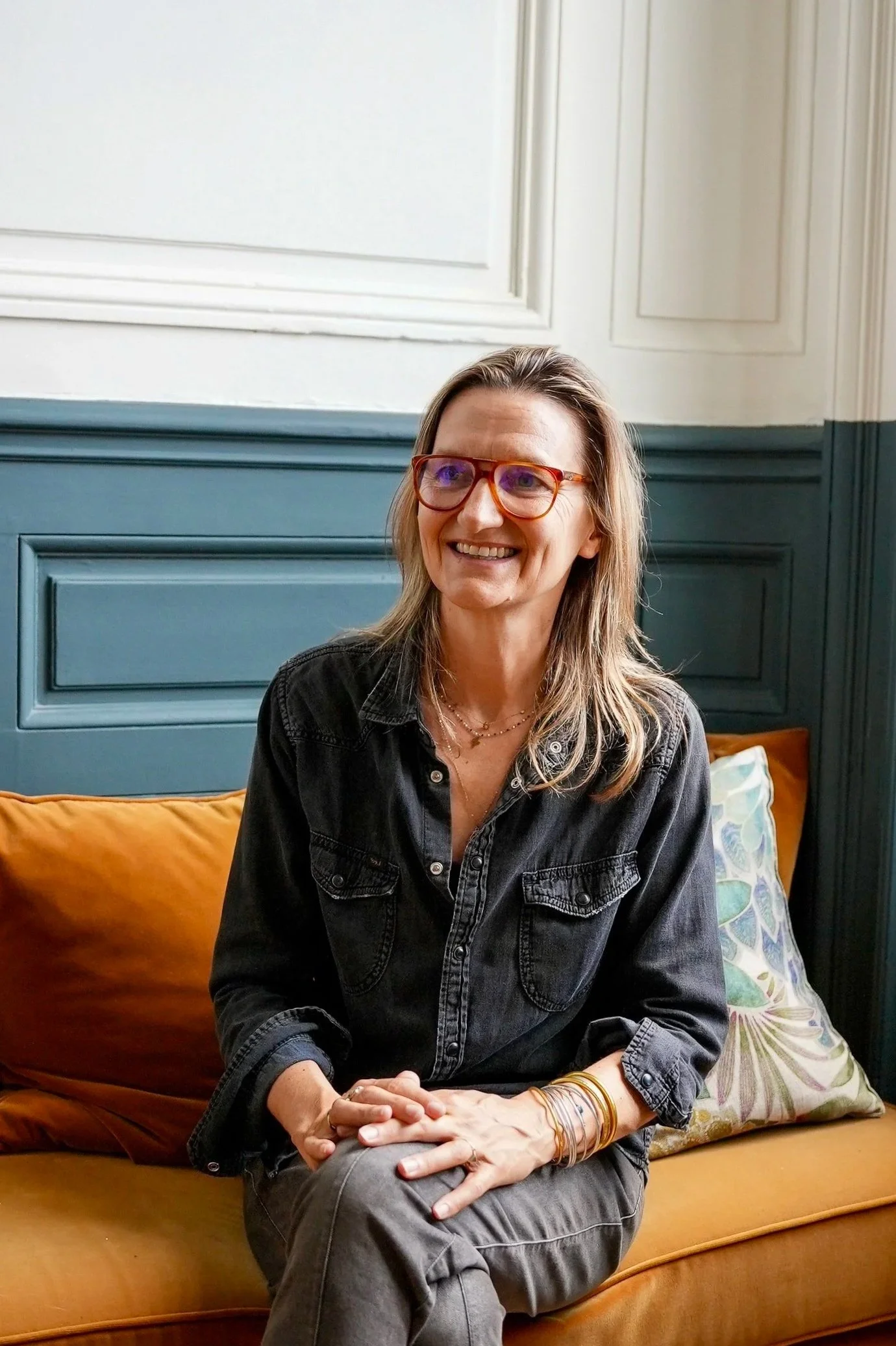 Une femme souriante avec des lunettes écailles, portant une veste en denim noir, assise sur un canapé en velours orange avec un coussin coloré à motifs, à l'intérieur d'une pièce avec des murs blanc et bleu, et un tableau blanc en arrière-plan.