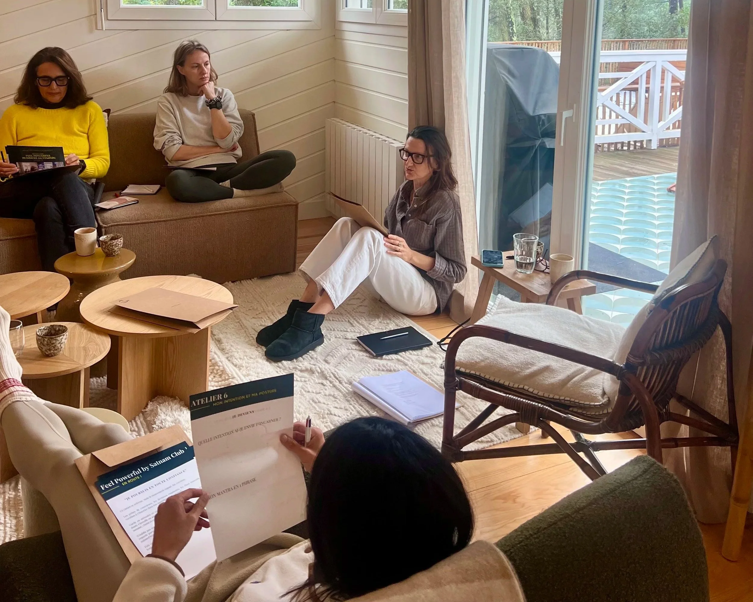 Groupe de quatre personnes assises dans une pièce, discutant et lisant des documents. La pièce a des murs en bois clair et une grande fenêtre avec vue sur une terrasse en extérieur.