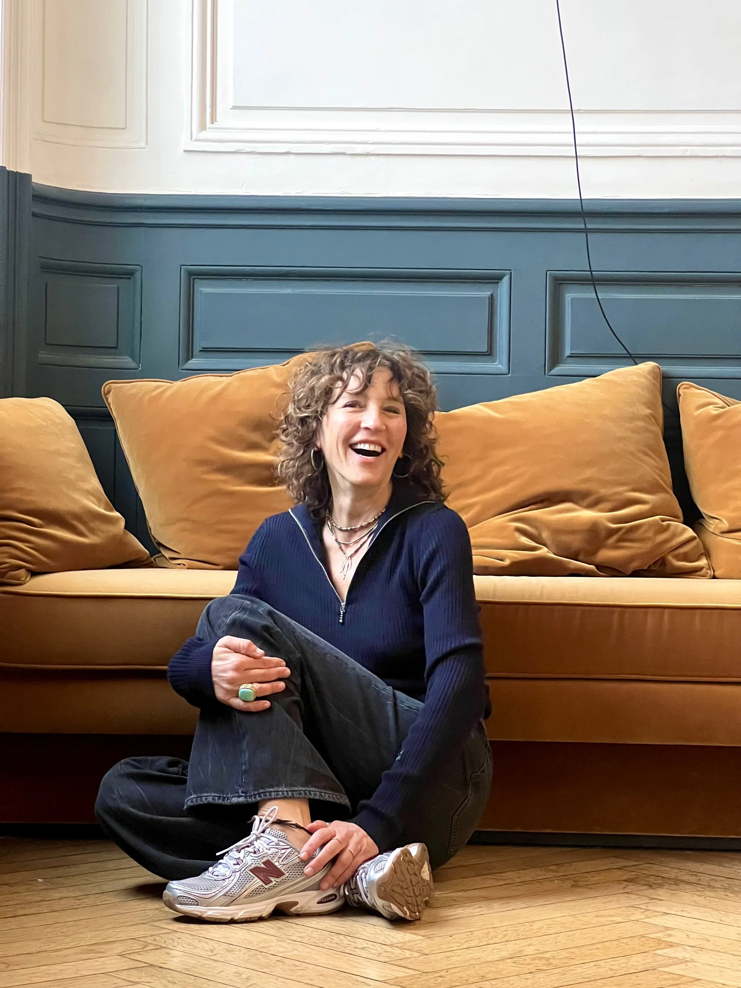 Une femme souriante avec des cheveux bouclés, assise par terre avec une jambe croisée, portant un sweat noir, des jeans noirs, des baskets gris et blanches, devant un canapé orange avec plusieurs coussins orange, dans un salon avec un mur bleu et un plancher en bois.