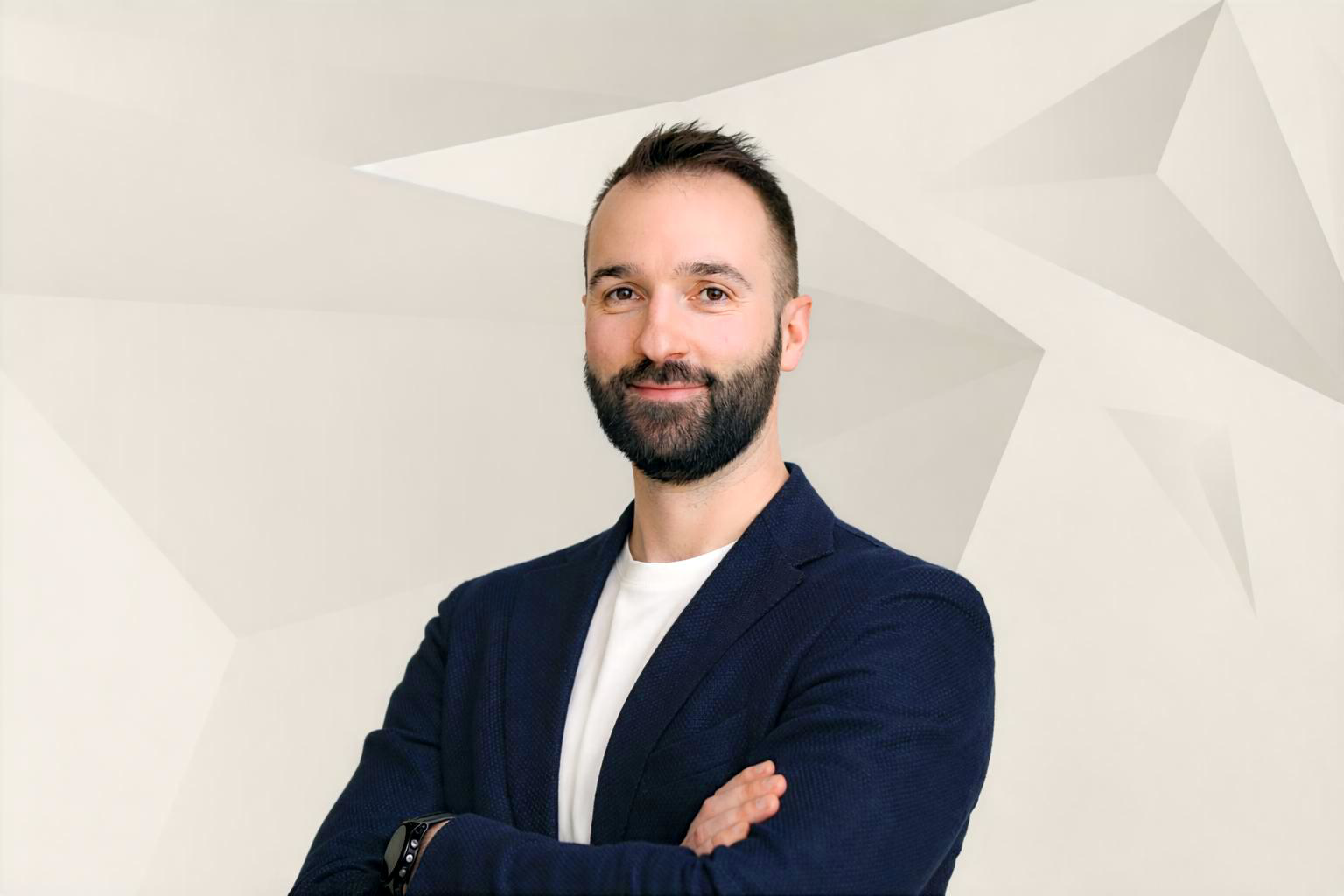 Ein Mann mit dunklem Haar und Bart trägt einen dunklen Blazer und ein weißes T-Shirt, steht vor einem weißen Hintergrund mit geometrischen Mustern, lächelt leicht und hat die Arme verschränkt.
