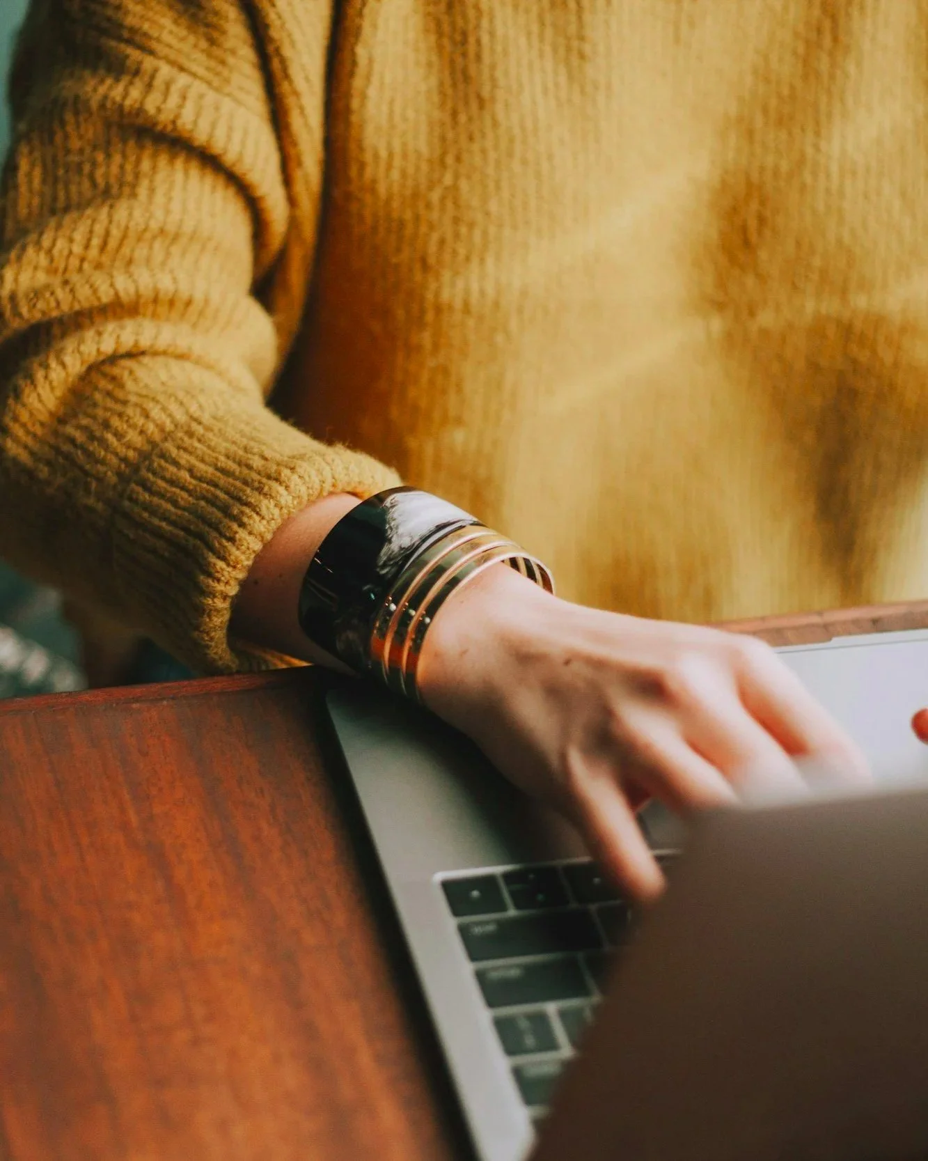Close-up of a person's hand with a decorative bracelet using a laptop computer at a wooden desk, wearing a yellow knitted sweater.