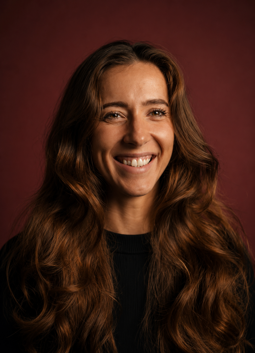 A woman with long, wavy brown hair and a big smile, wearing a black top, against a dark red background.