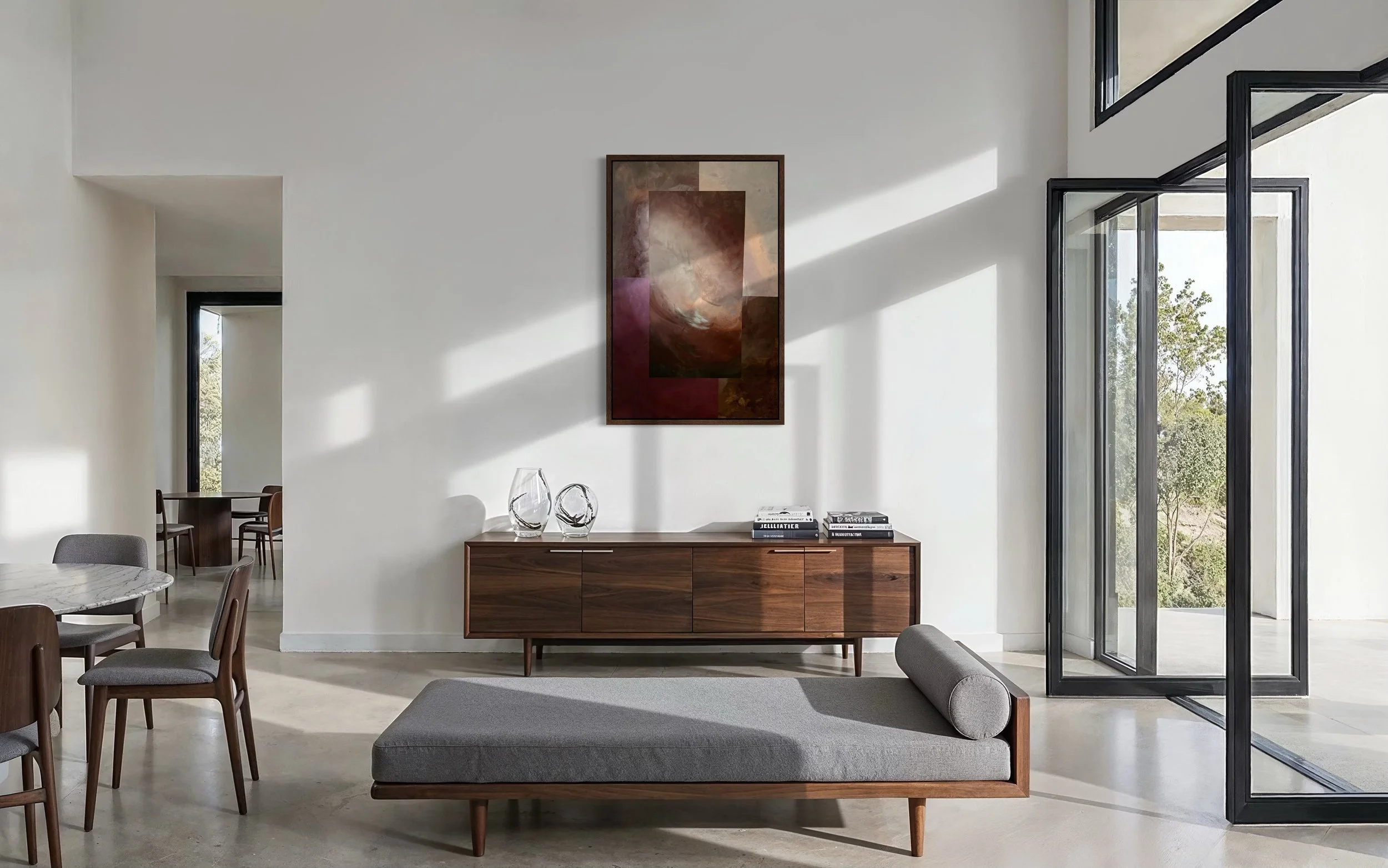 Modern living room with white walls, large windows, a wooden sideboard with decorative glass objects and books, and a gray ottoman with a cylindrical pillow, sunlight streaming in.